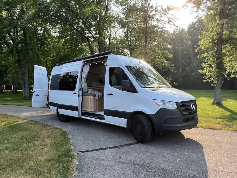 A white Mercedes-Benz Sprinter van parked on a driveway in a park-like setting with trees and grass, with the side door open showing a well-equipped interior.