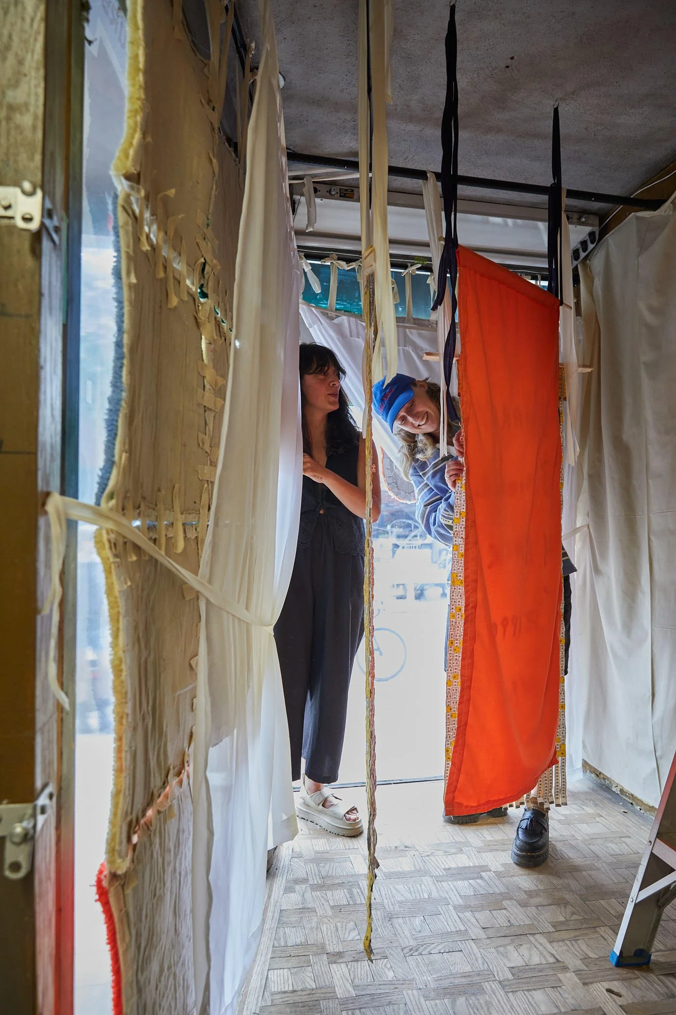 Shannon Stride. Two women peek through a gap in a fabric curtain display inside a booth with hanging textiles.