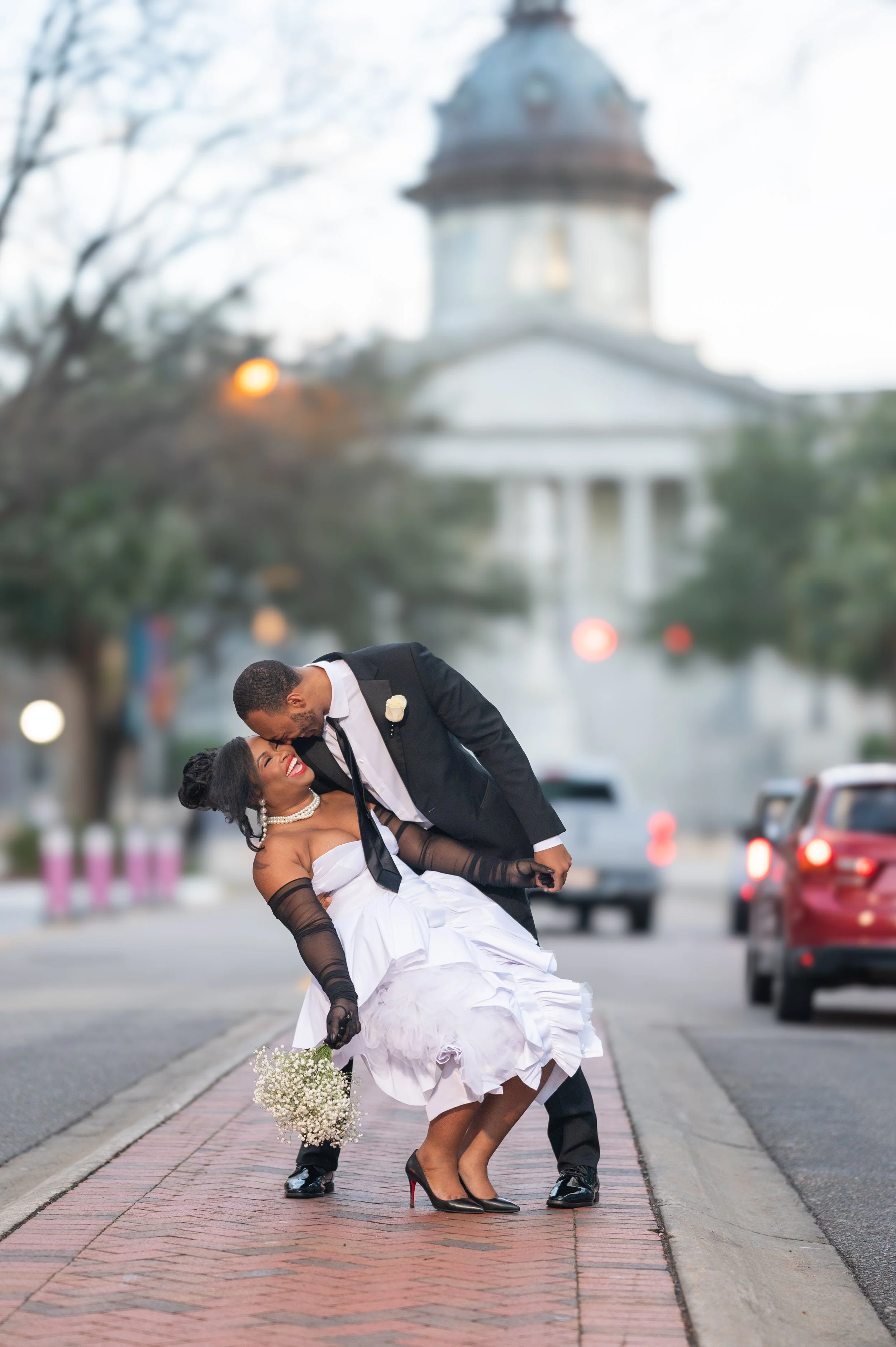 A newlywed couple dancing on a city sidewalk with a government building in the background. The bride wears a white dress, black gloves, and high heels, and holds a bouquet of white flowers. The groom wears a black suit and white shirt. They are smiling and enjoying their moment together.