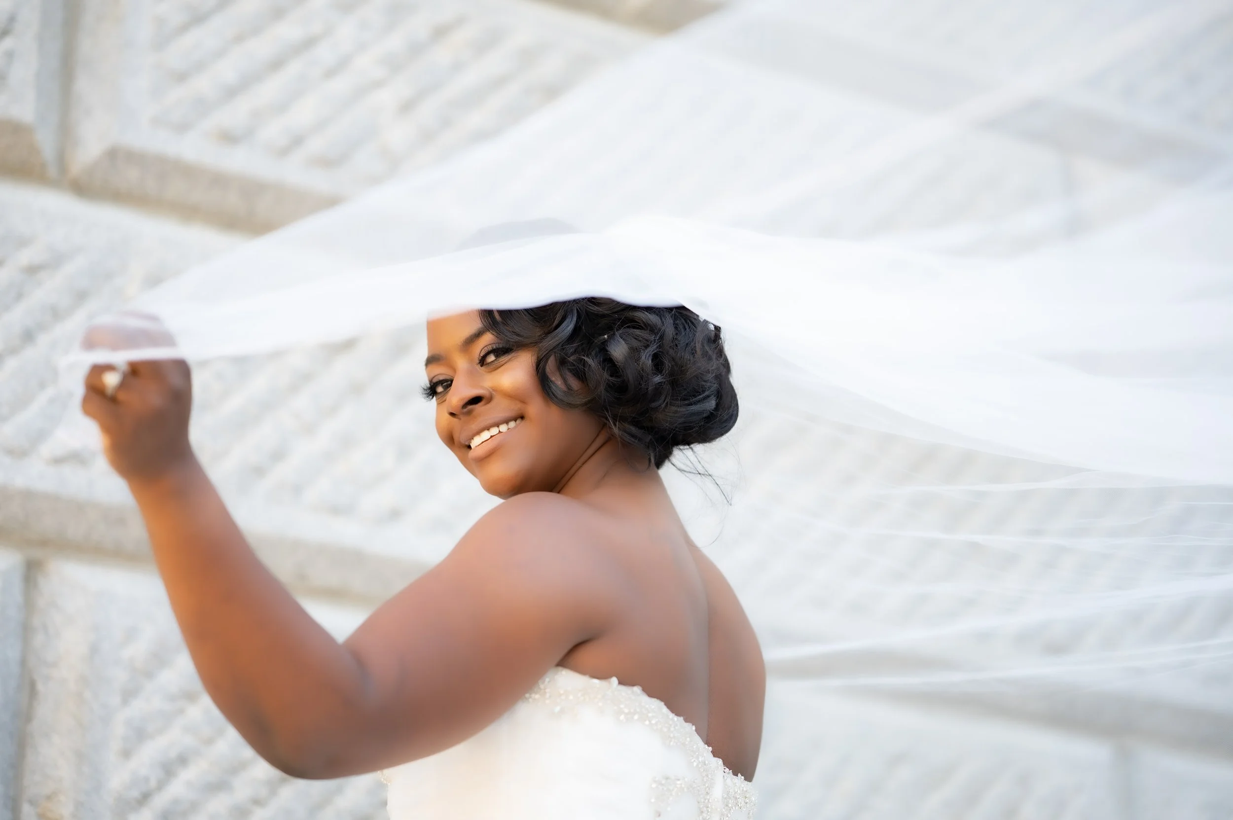 Smiling bride in a white wedding dress holding her veil outdoors near stone steps.