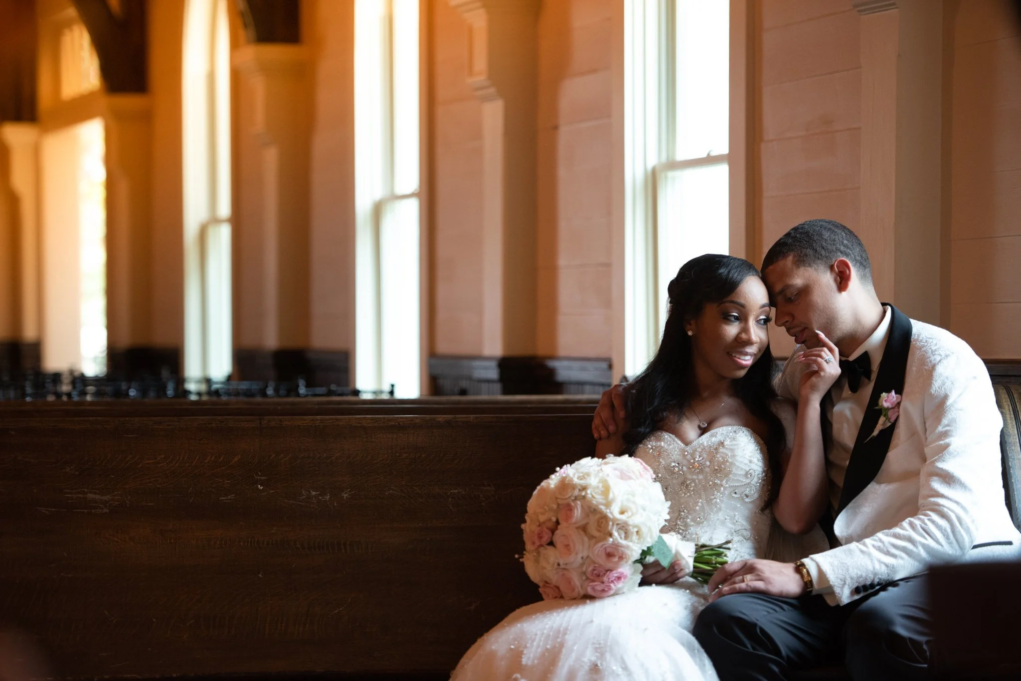 A bride and groom sitting close together on a bench in a church, with the bride holding a bouquet of pink and white roses, both dressed in wedding attire and sharing an intimate moment.