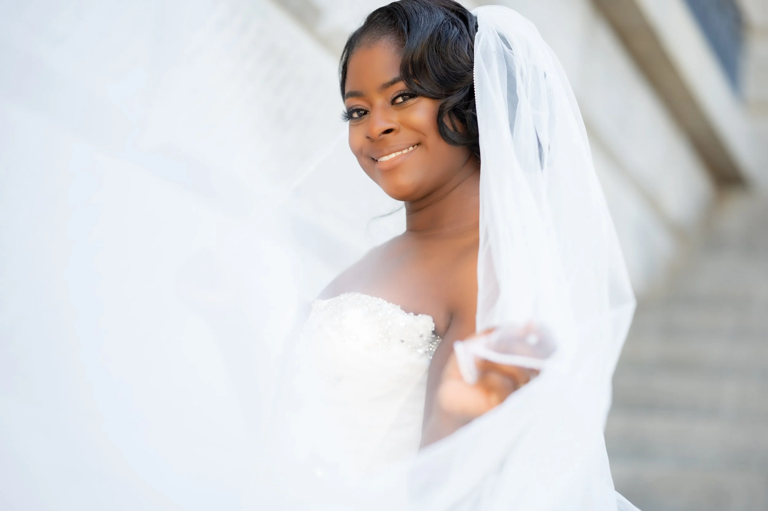 A bride smiling, wearing a white wedding dress and veil, holding the edge of her veil, outdoors with stairs in the background.