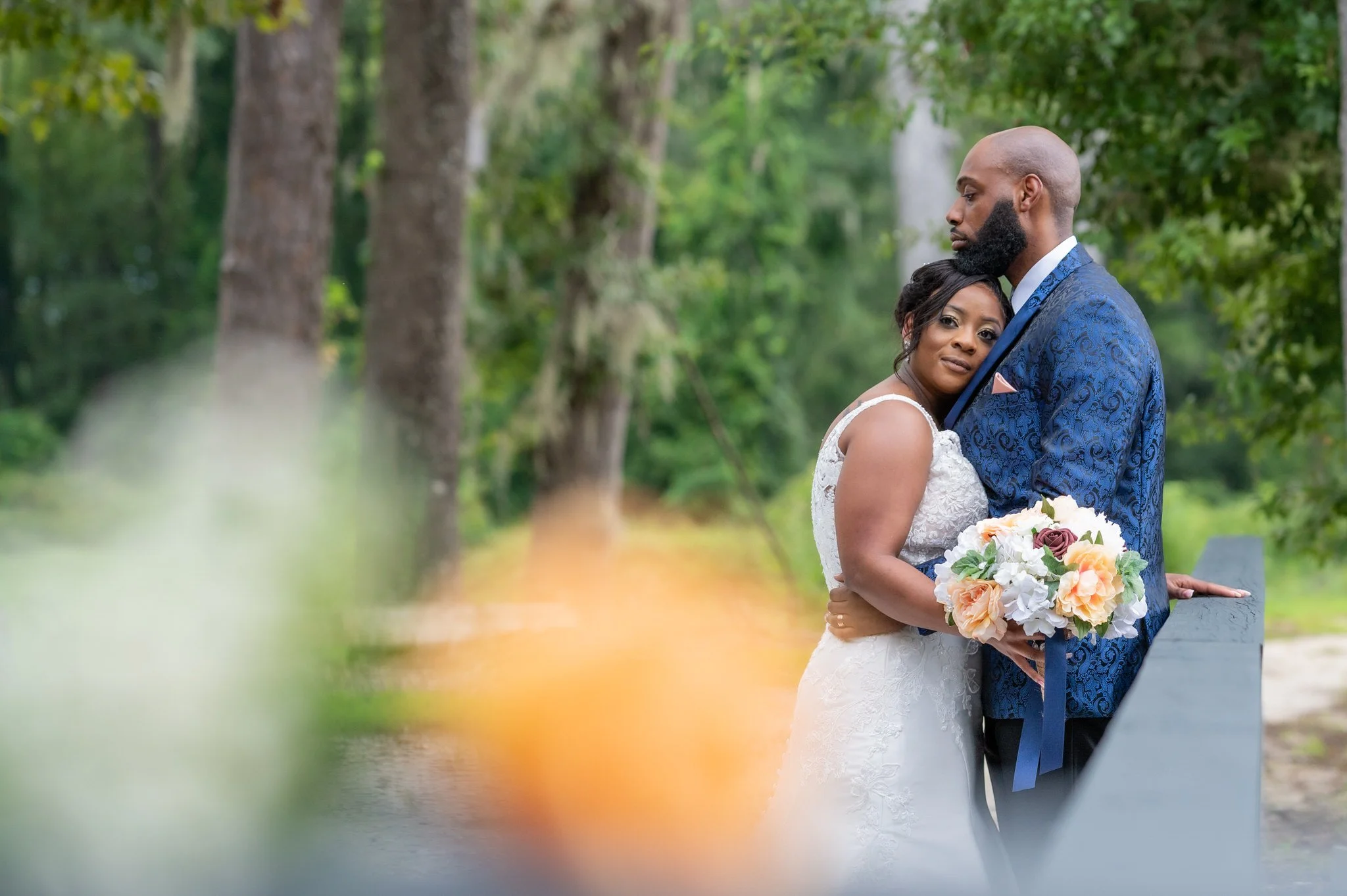 A bride in a white lace wedding dress and a groom in a blue patterned suit stand closely together outdoors on a green wooded path, with the bride holding a bouquet of white and peach flowers. The bride leans her head on the groom's chest as he looks contemplative.