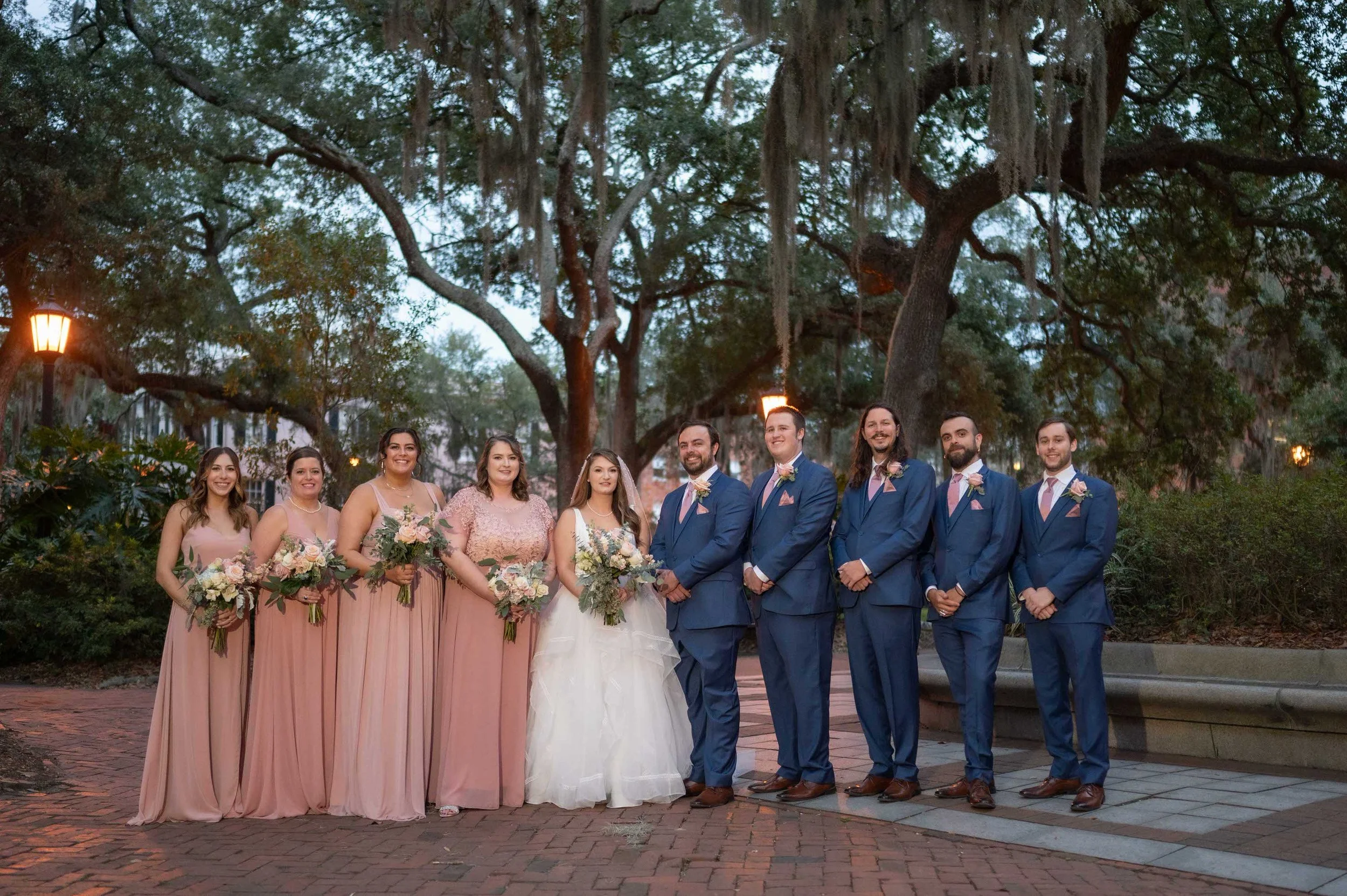 Wedding party standing outdoors. Bridesmaids in pink dresses and groomsmen in blue suits with pink ties. Bride and groom in the center. Large trees with hanging moss in the background, evening setting with warm streetlights.