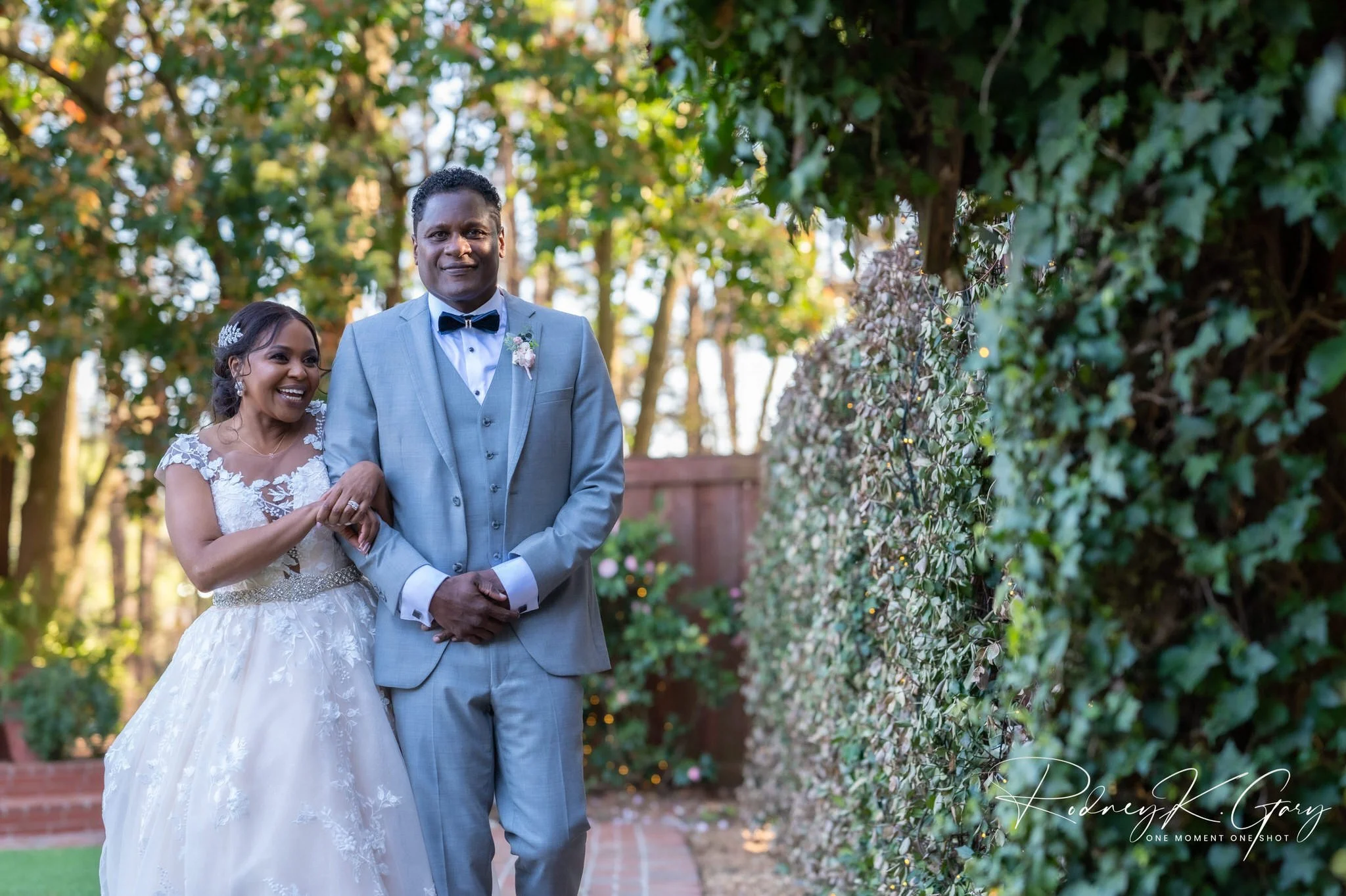 A bride and groom holding hands and walking outdoors, surrounded by greenery and trees, on their wedding day.