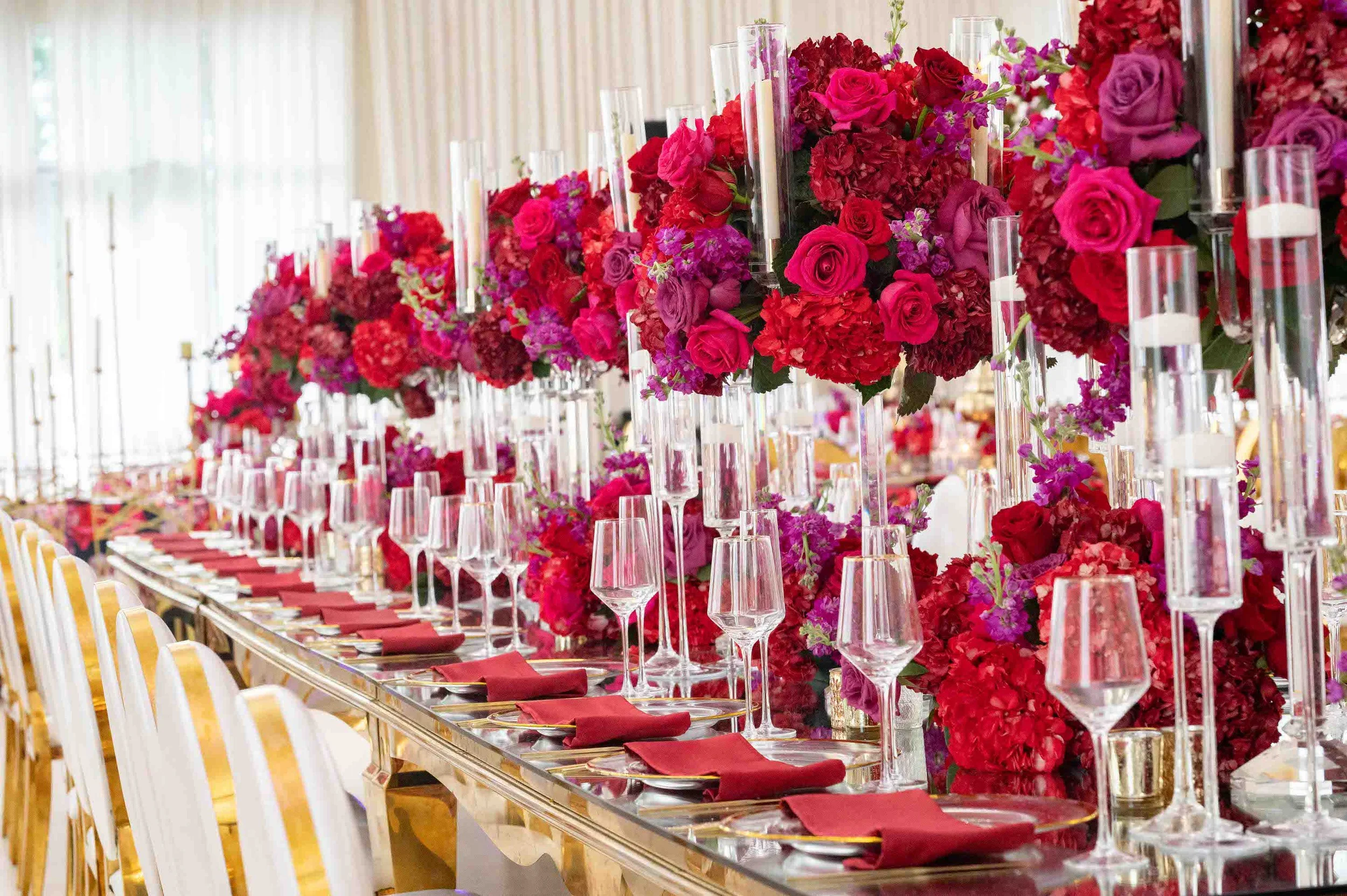 Long banquet table decorated with tall floral centerpieces of pink and red flowers, clear candles, wine glasses, and red napkins arranged on plates, set for a fancy event.