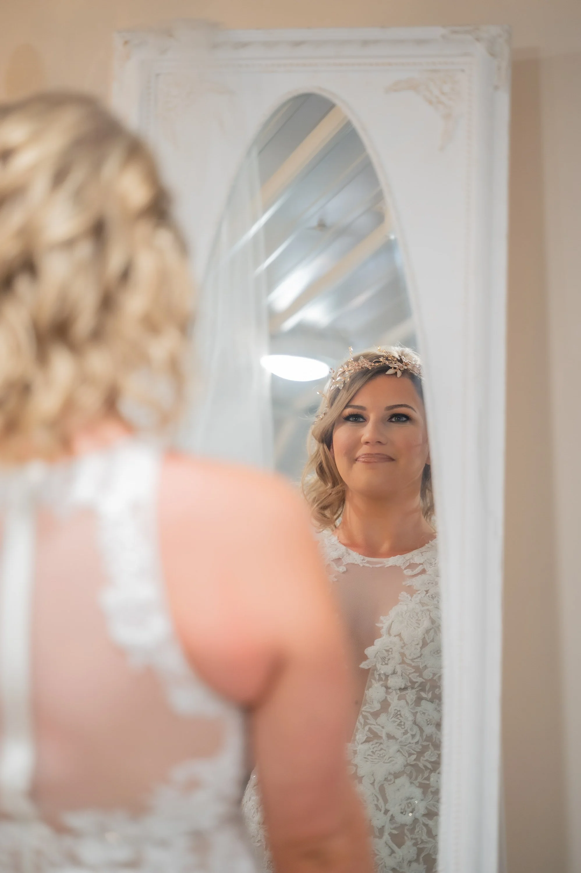 Bride looking at herself in a mirror, wearing a lace wedding dress and a floral headpiece, in a softly lit room.