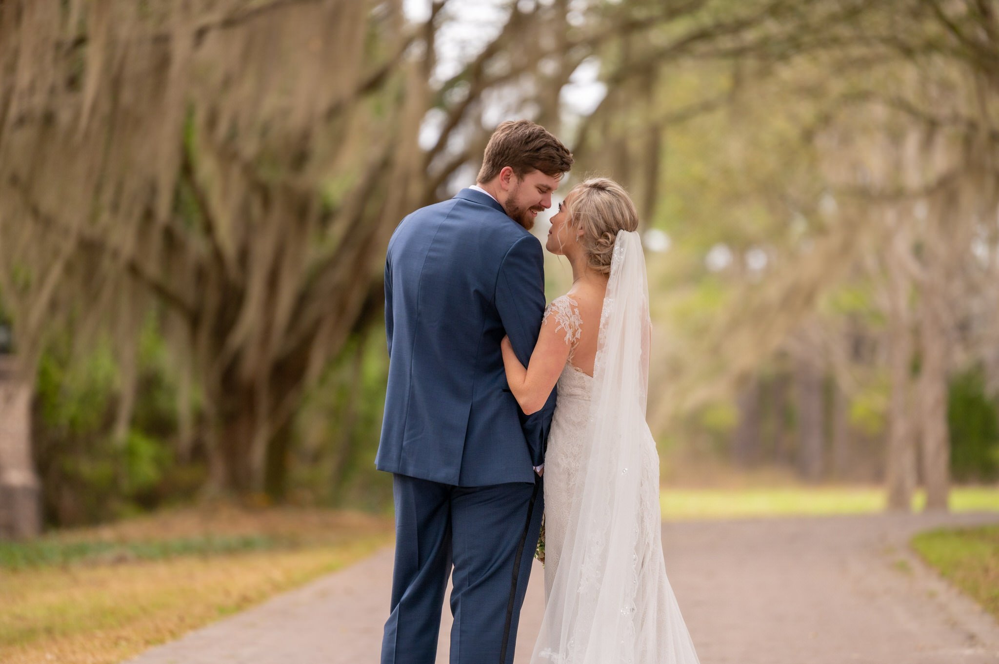 A bride and groom stand close on a tree-lined dirt path, sharing an intimate moment at their wedding, the bride wearing a white lace gown and veil, the groom in a blue suit, surrounded by green trees.
