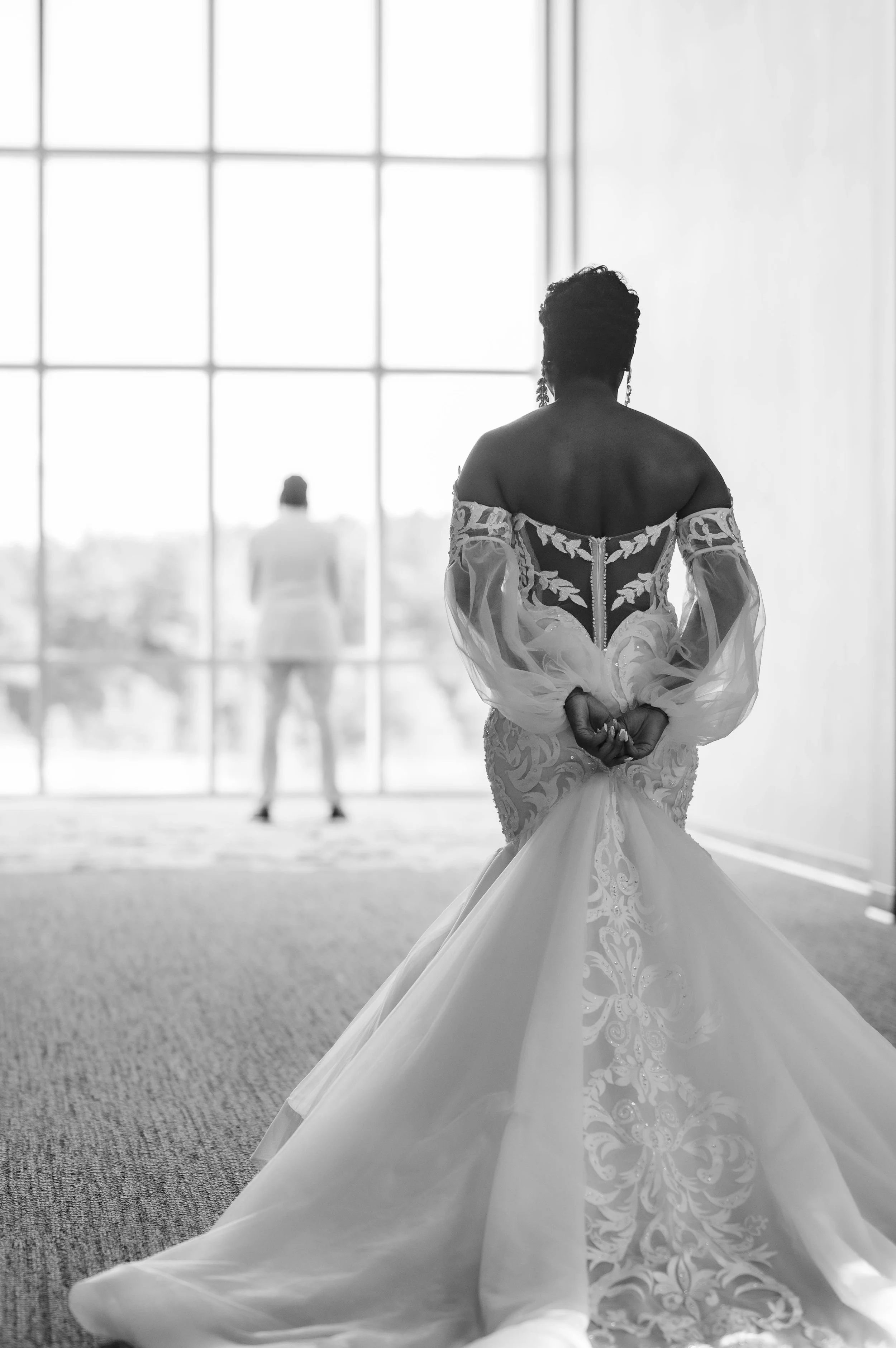 Brides wearing wedding dresses, standing by large windows in a sunlit room