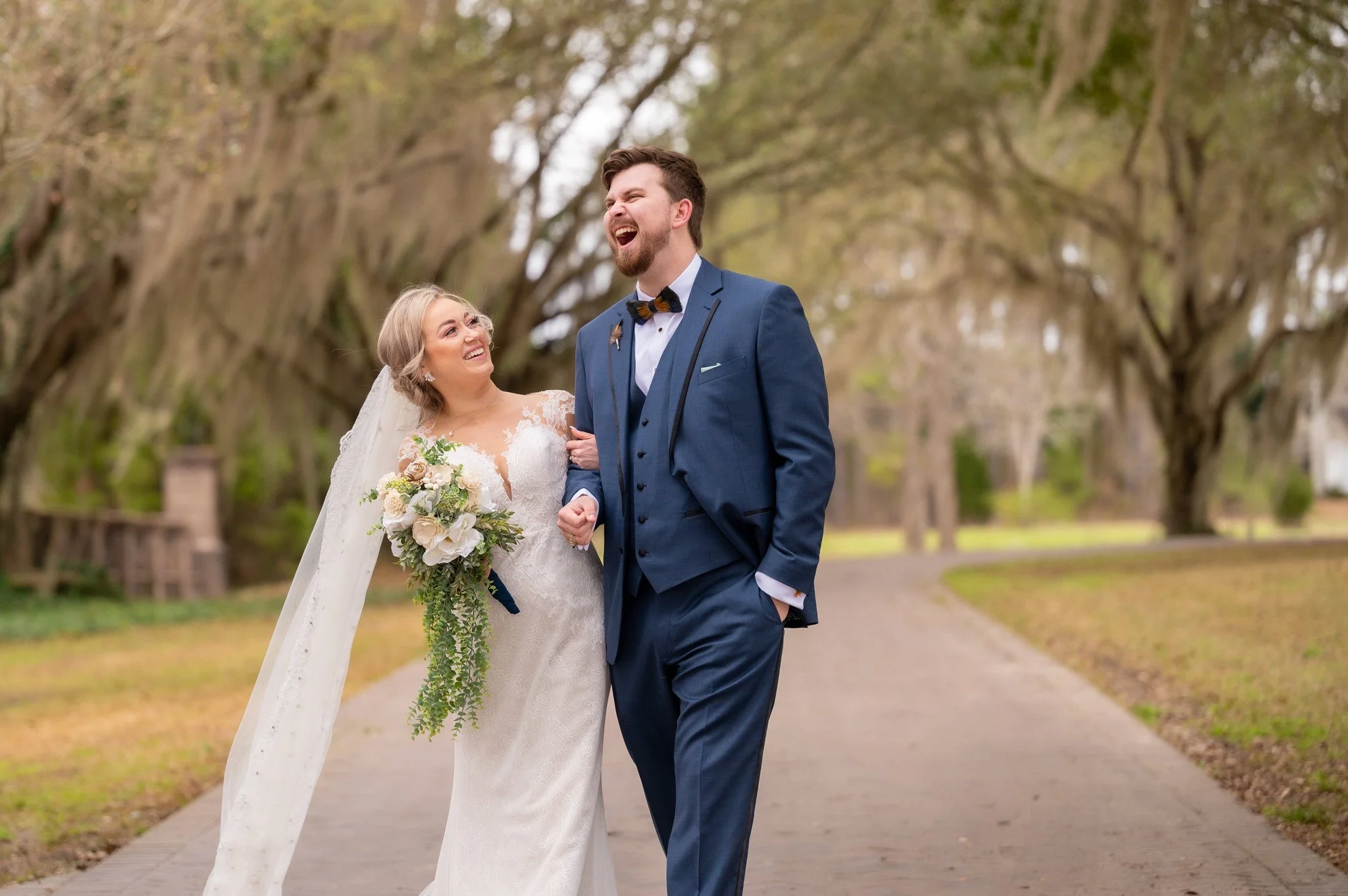 A bride and groom walking together outdoors, smiling and laughing under a canopy of trees, during a wedding photoshoot.