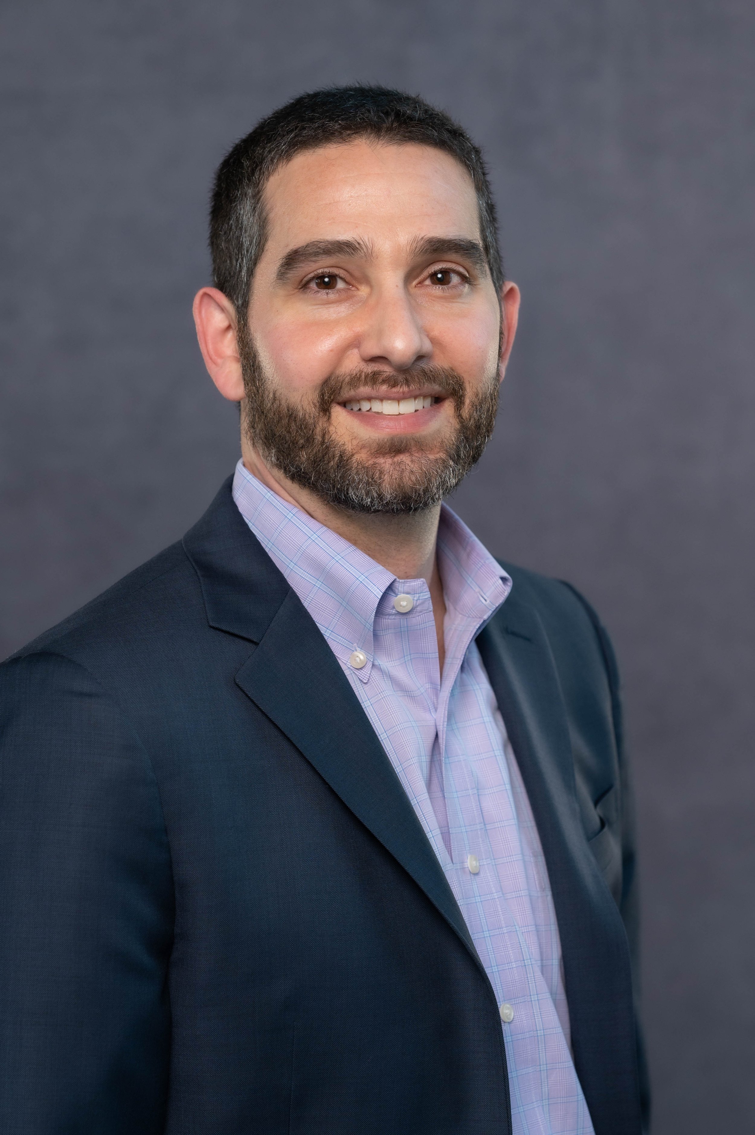Headshot of a man with short dark hair and a beard, wearing a dark blazer and light purple collared shirt, smiling against a gray background.