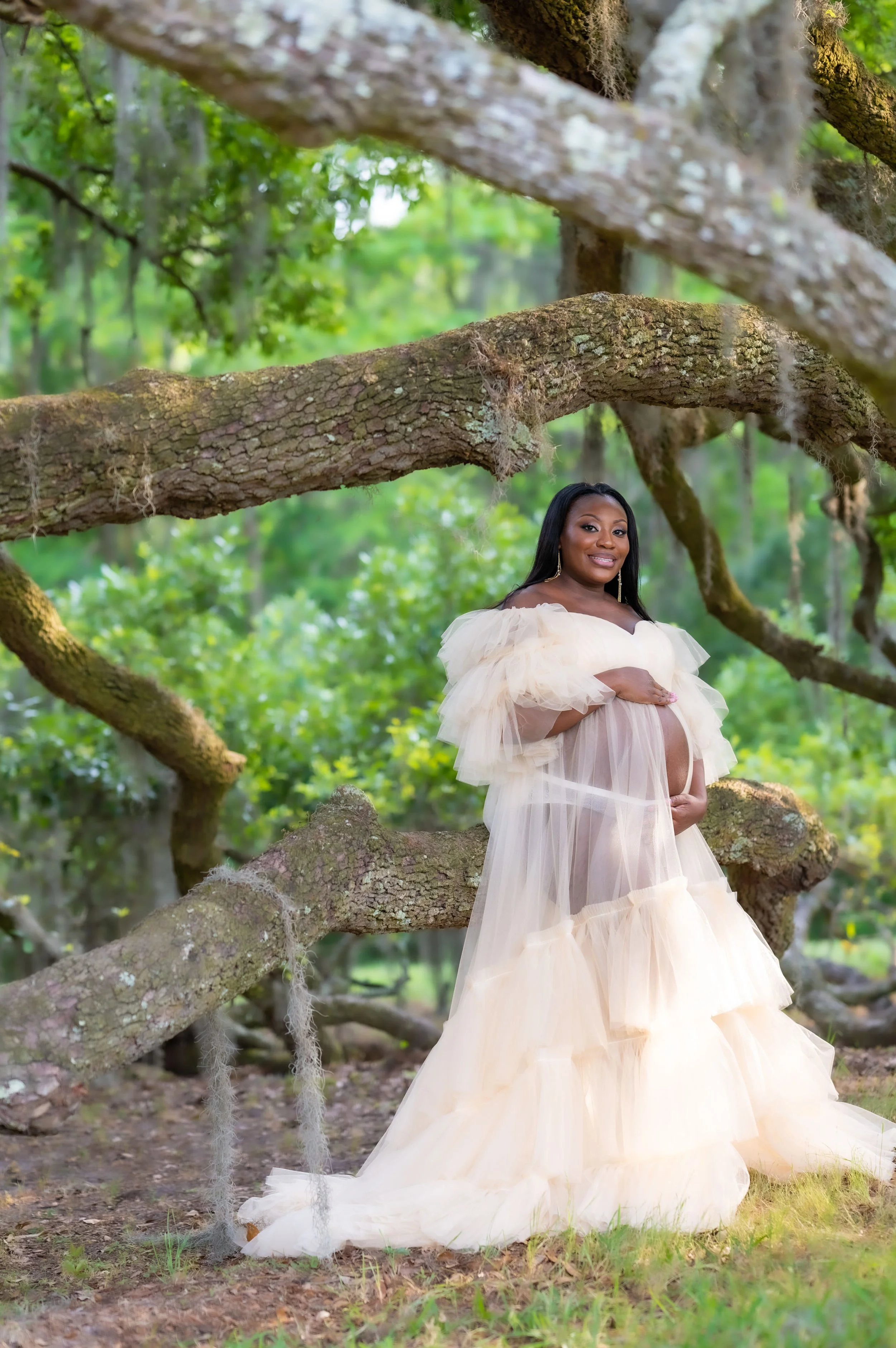 Pregnant woman in a flowing beige dress standing in a green forest, holding her belly and smiling at the camera.