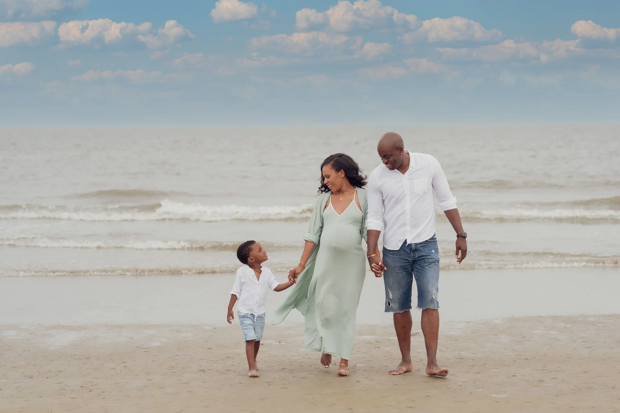 Family of three walking hand in hand on a beach with ocean and cloudy sky in the background.
