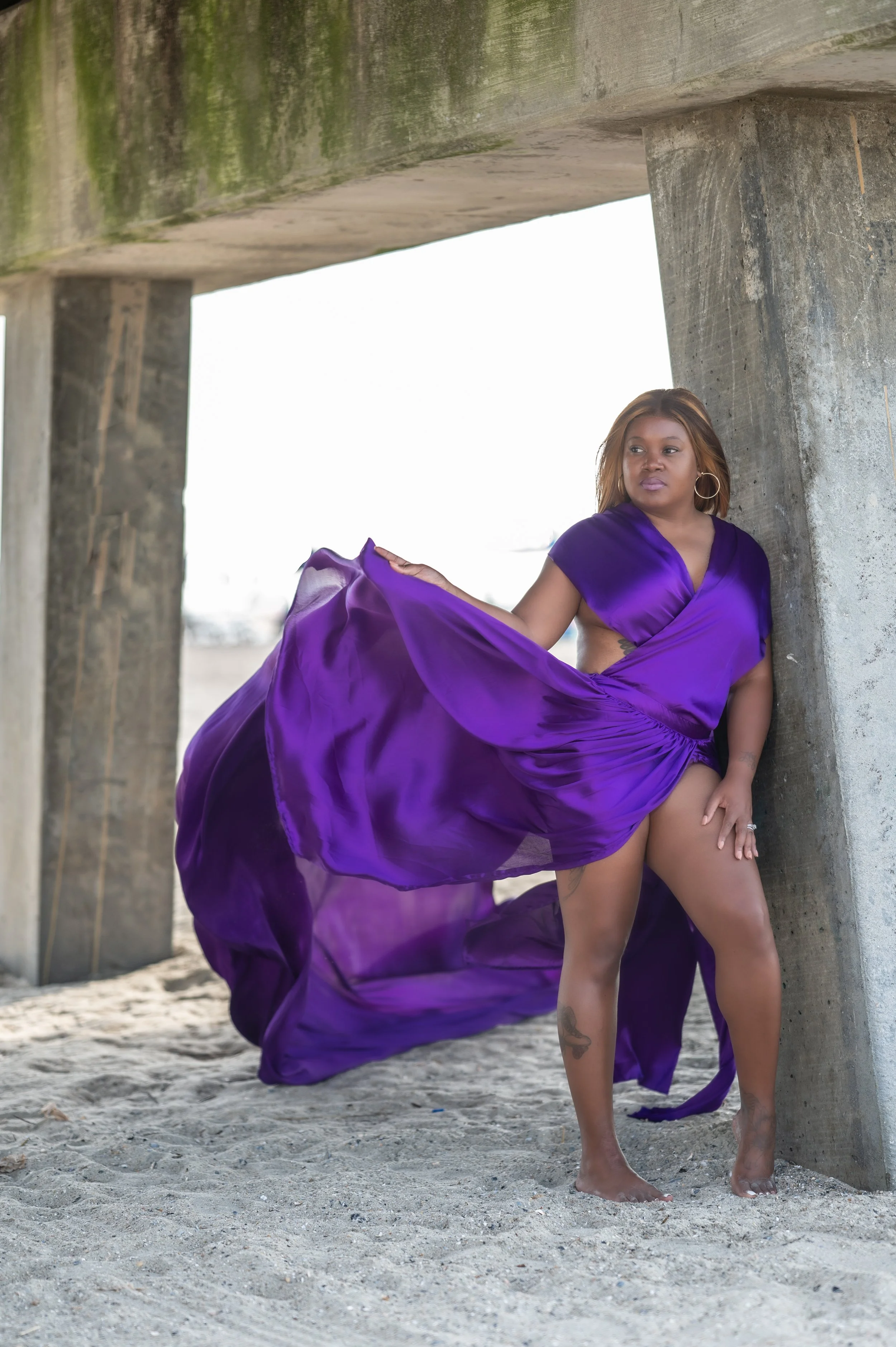 A woman in a flowing purple dress standing barefoot on sandy ground under a concrete structure