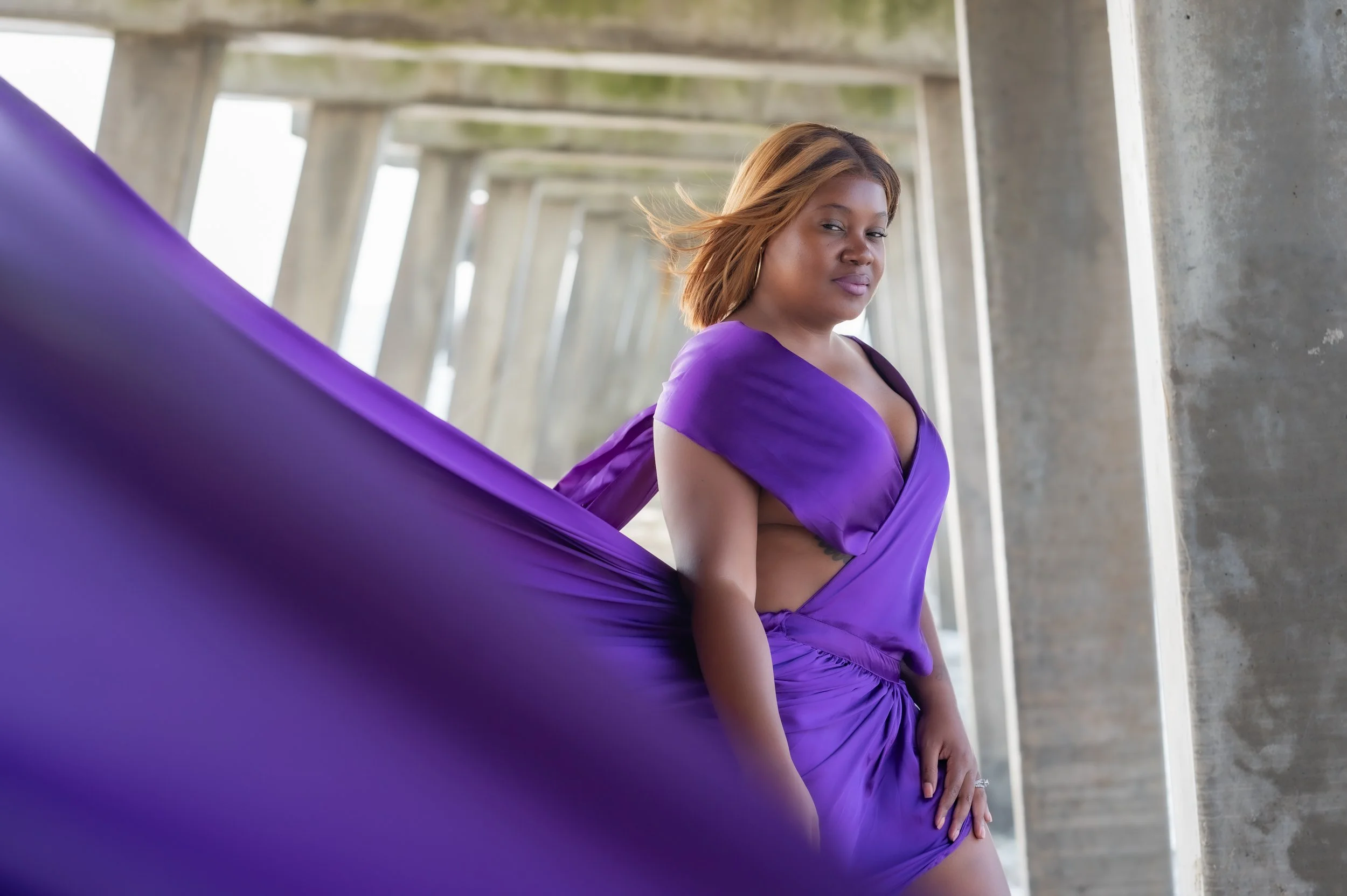 Woman in a purple dress standing under a bridge with concrete pillars.