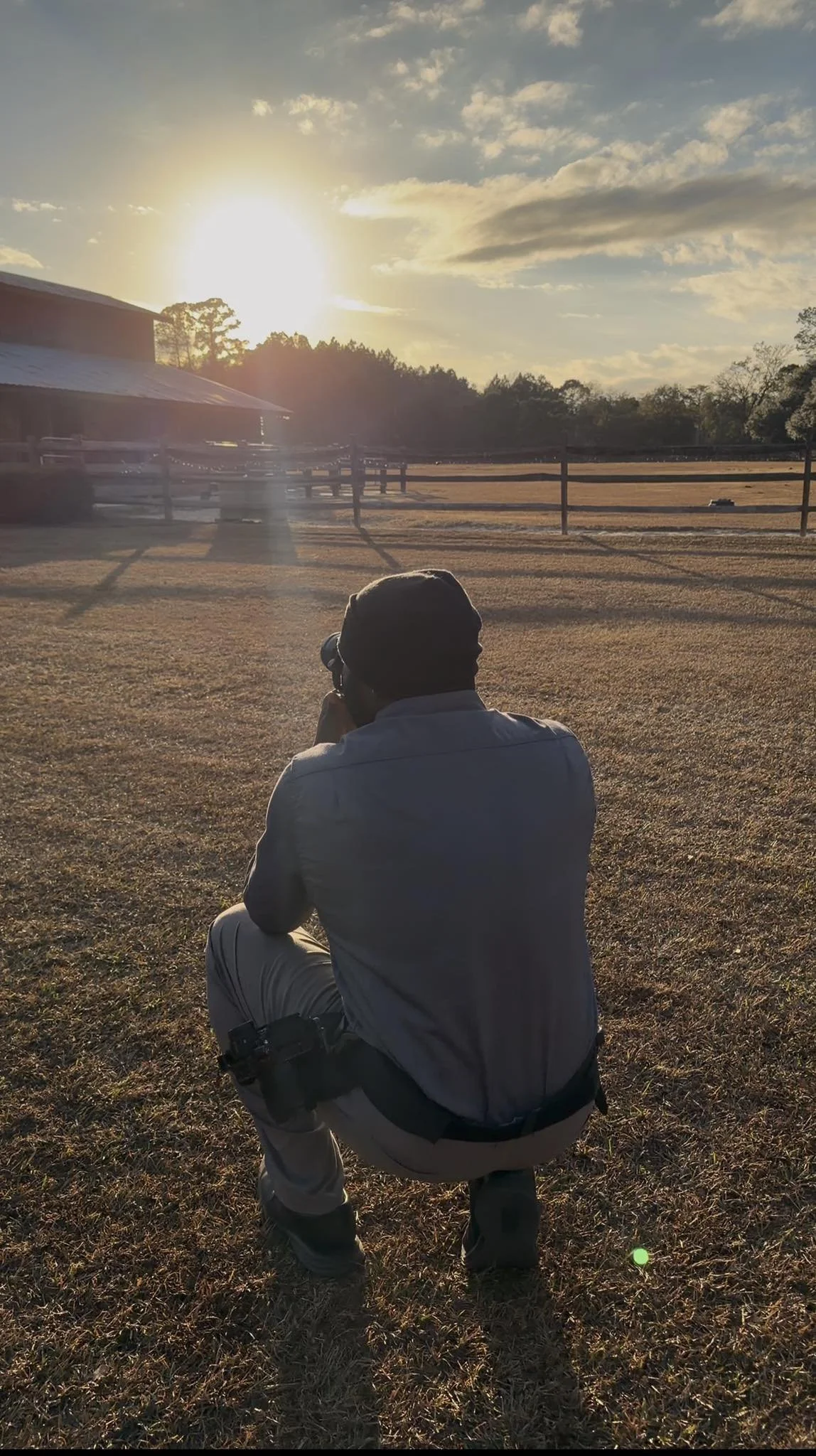 Photographer crouches on dry grass field, aiming camera at sunset sky with clouds, nearby structure, and a fence in the background.