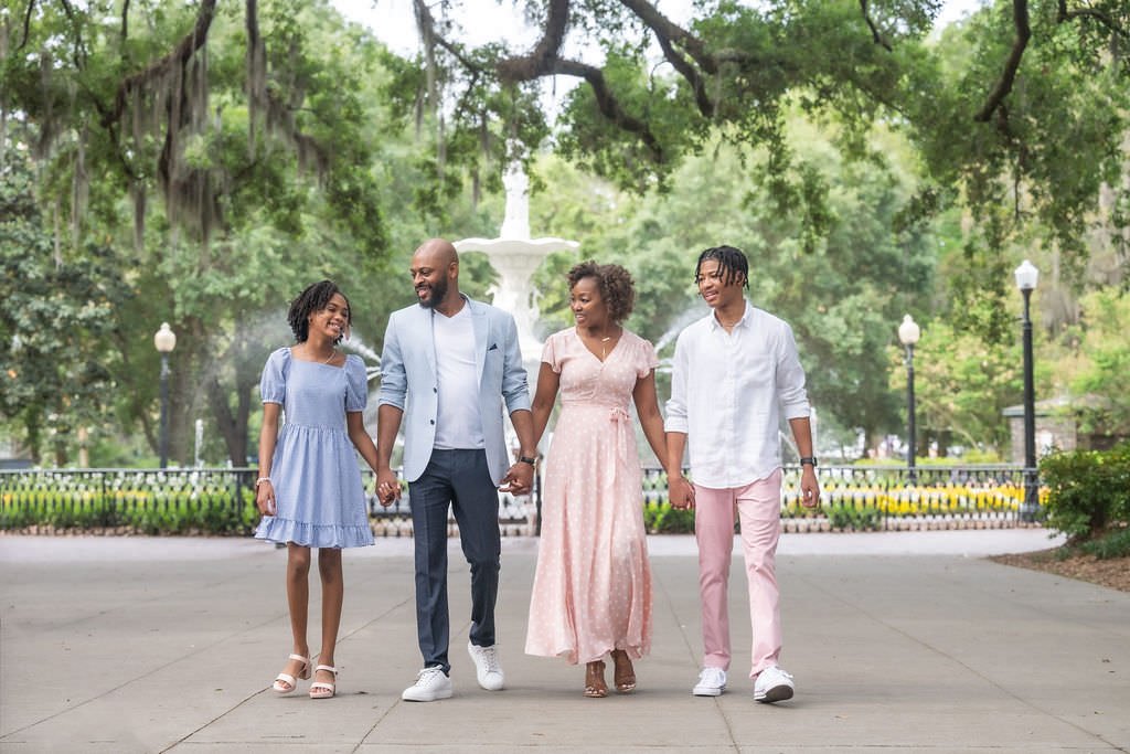 Family walking hand in hand in a park with large trees, a fountain, and lampposts in the background.