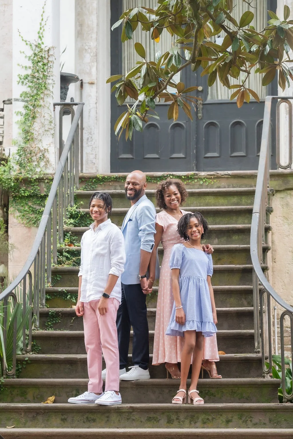 A family of four smiling and holding hands on the steps of a building front, with a large leafy tree overhead.