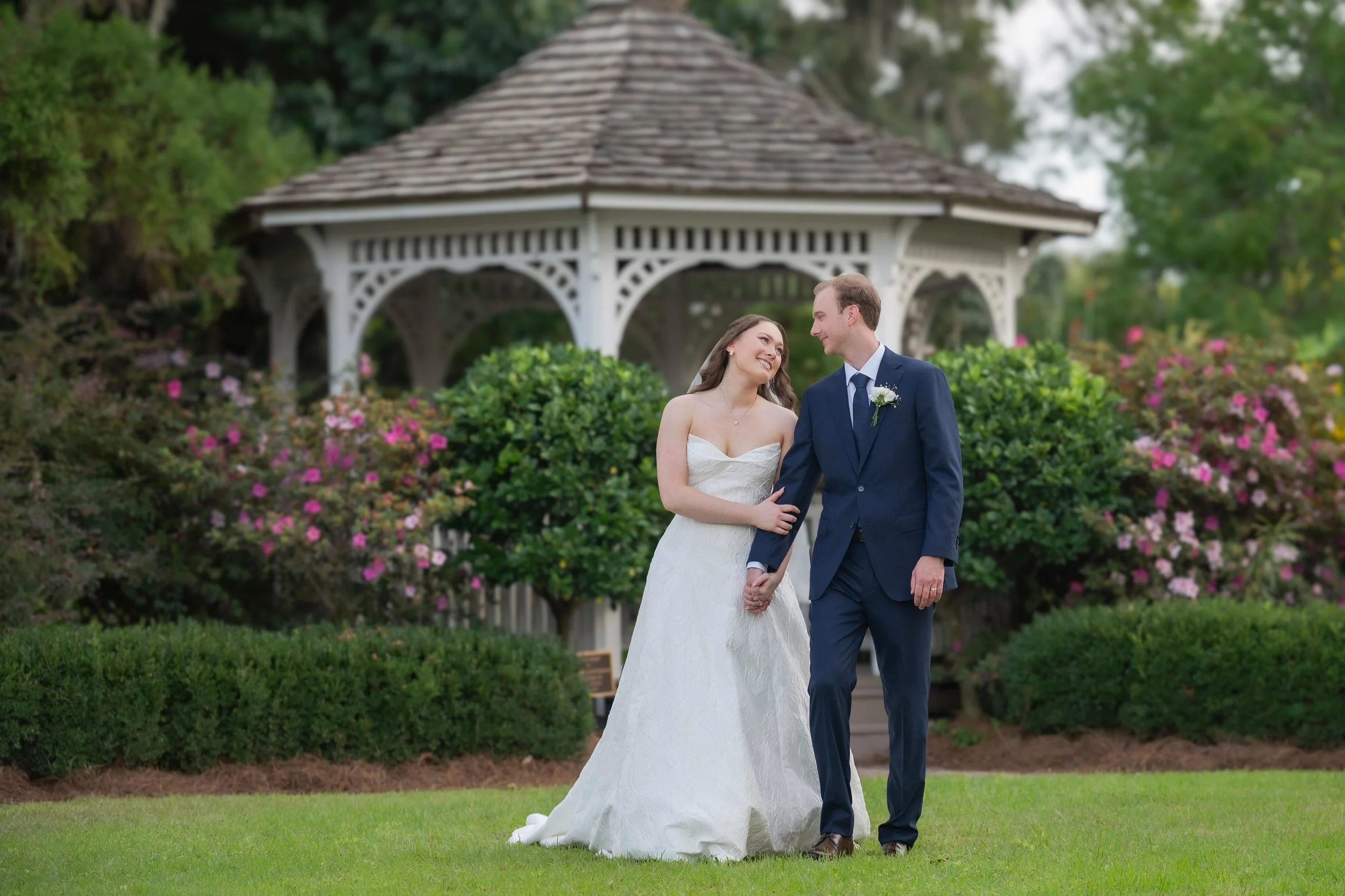 A bride and groom walking hand in hand on a lawn, smiling at each other, with a gazebo and pink flowering bushes in the background.