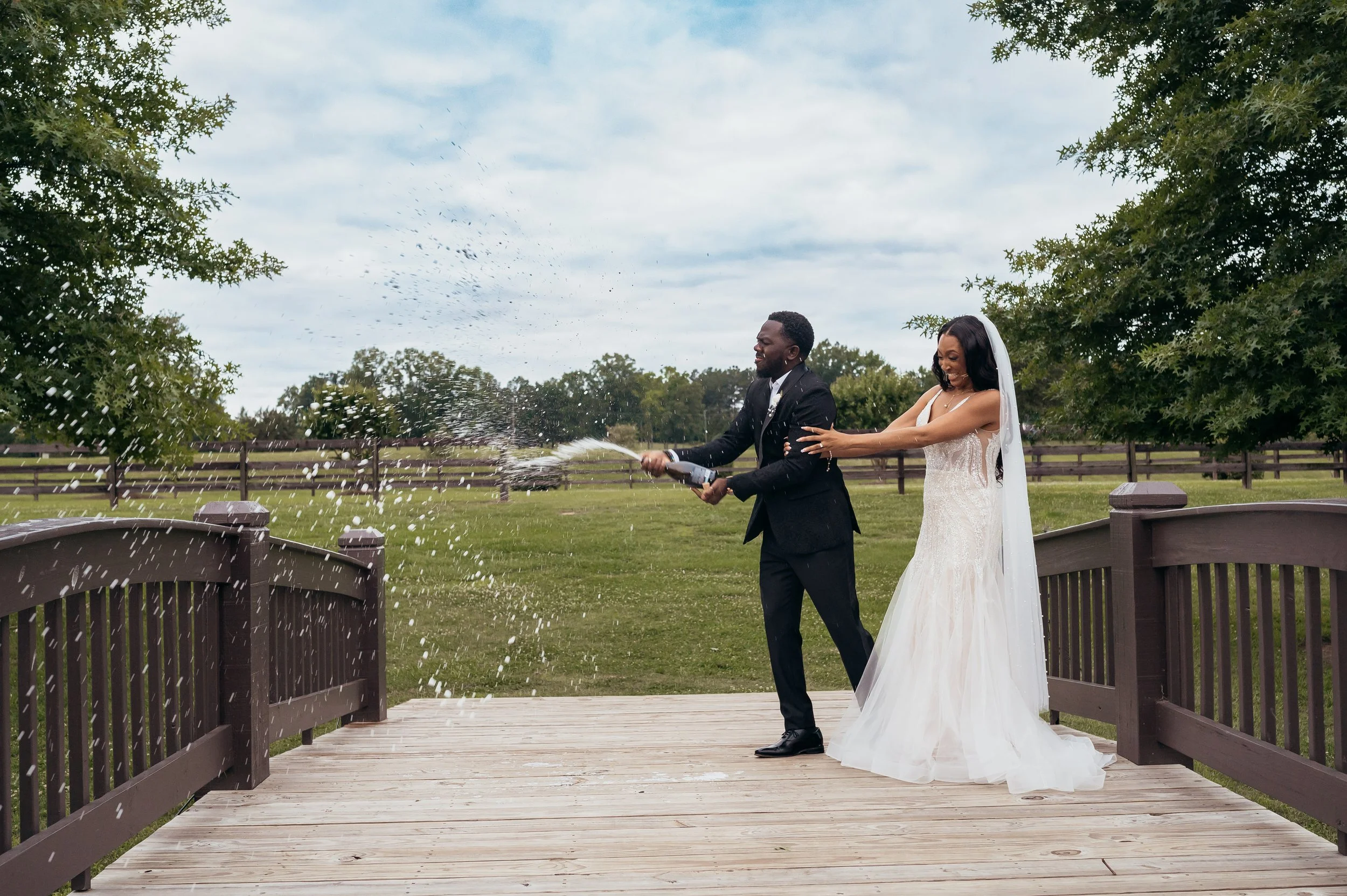 A newlywed couple on a wooden bridge celebrating by spraying champagne outdoors with trees and open sky in the background.