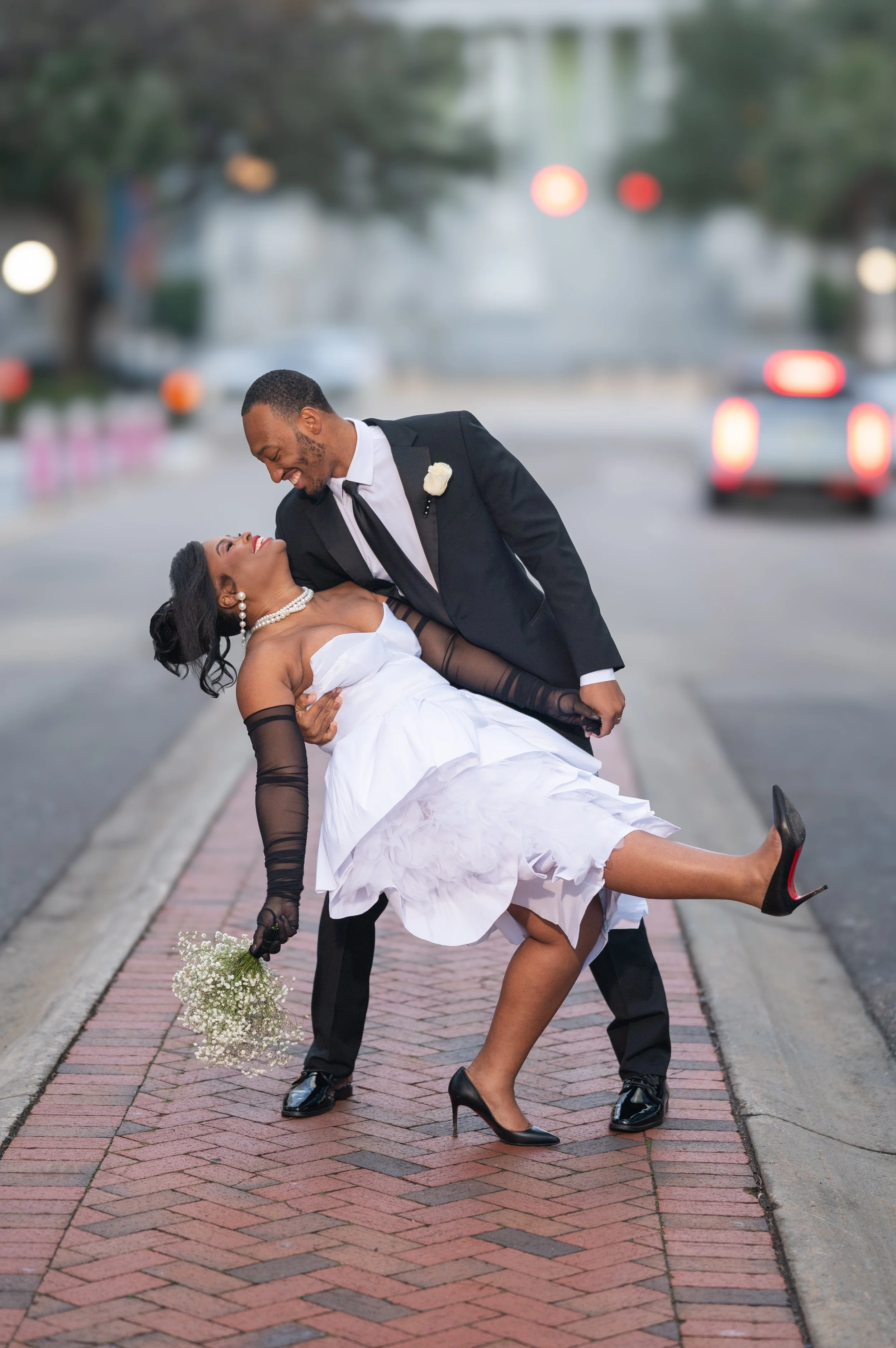 A newlywed couple dancing outside on a city sidewalk during the evening. The bride, in a white dress and black sheer gloves, is being dipped by the groom, who is wearing a black suit with a white shirt and black tie. She is holding a small bouquet of