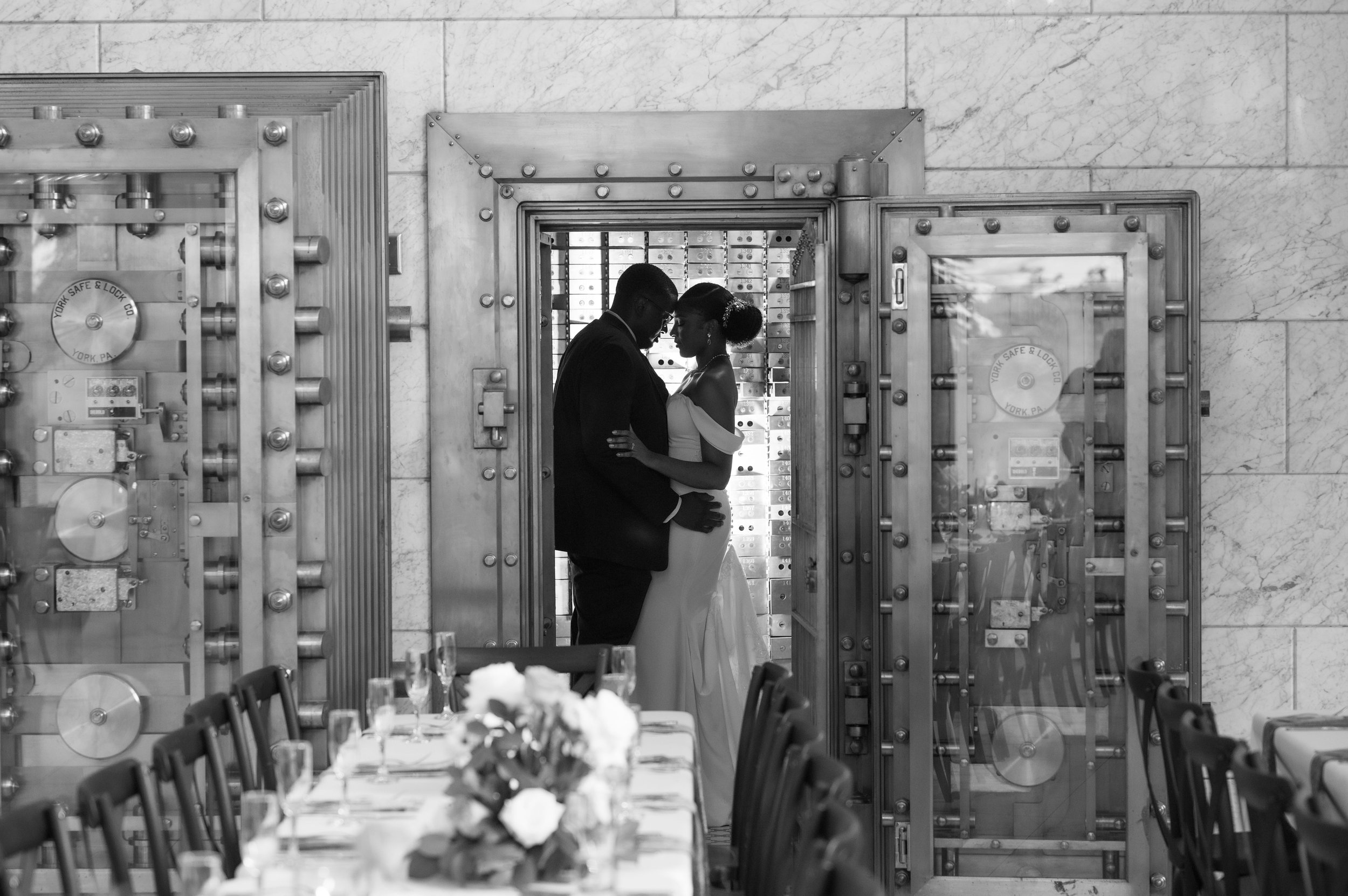 A black and white photo of a couple in wedding attire standing in a doorway of a bank vault. They are facing each other, close together, with the woman's arms around the man's neck and the man's hands on the woman's waist. The foreground shows a tabl