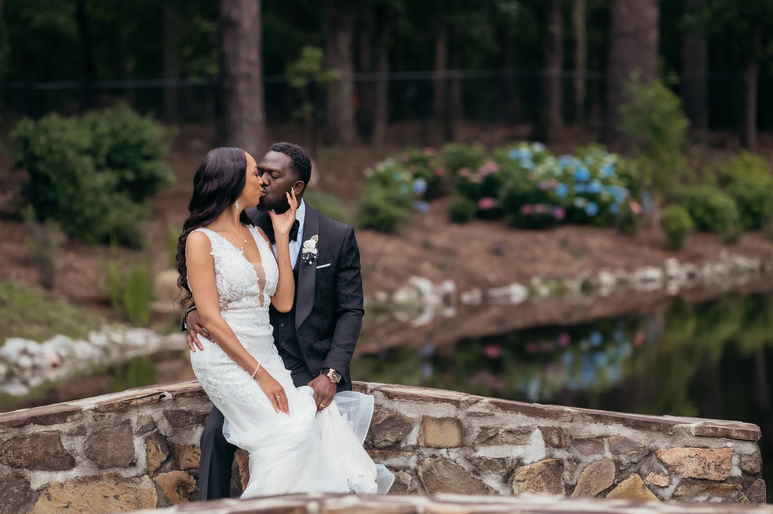 Couple in wedding attire sharing a kiss by a small pond with trees and flowering bushes in the background.