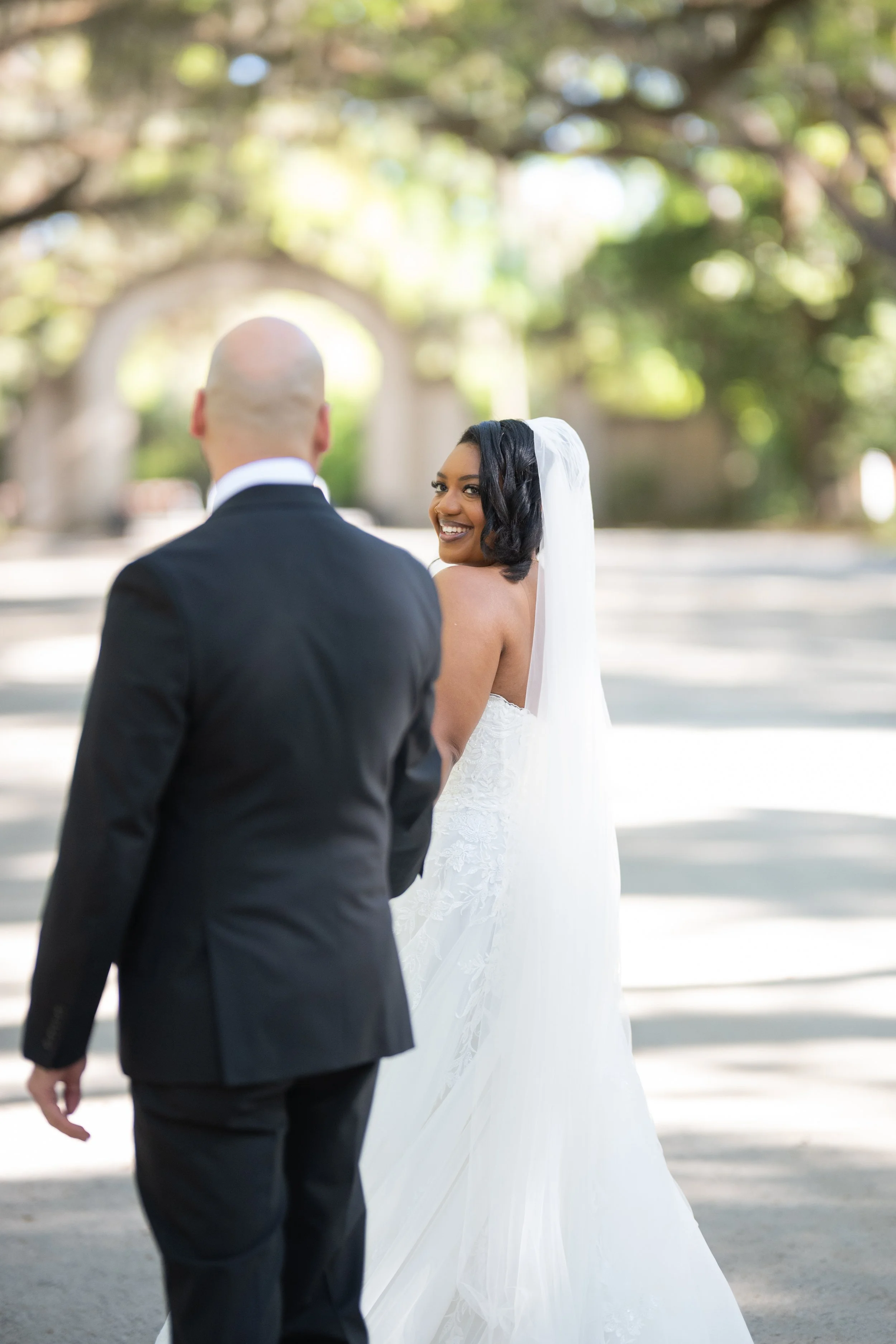 A bride in a white wedding dress with a veil smiles at the groom, who is dressed in a black suit, outdoors under a tree with sunlight filtering through the leaves.