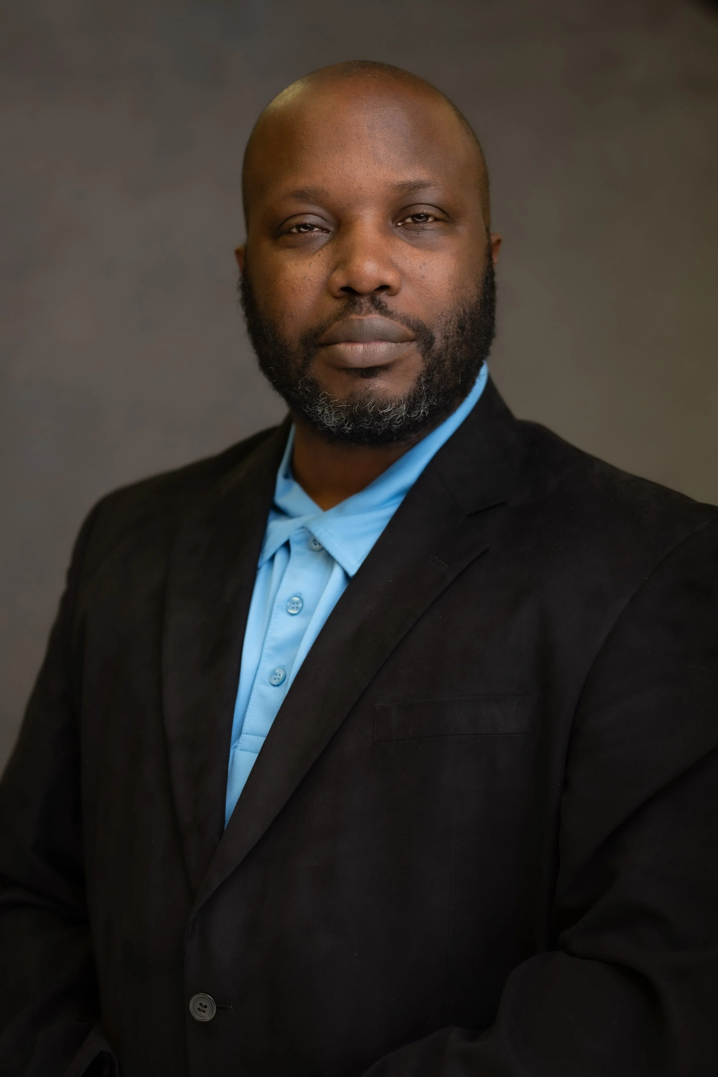 A man with a beard and bald head wearing a black suit jacket and a light blue collared shirt, looking directly at the camera against a neutral background.