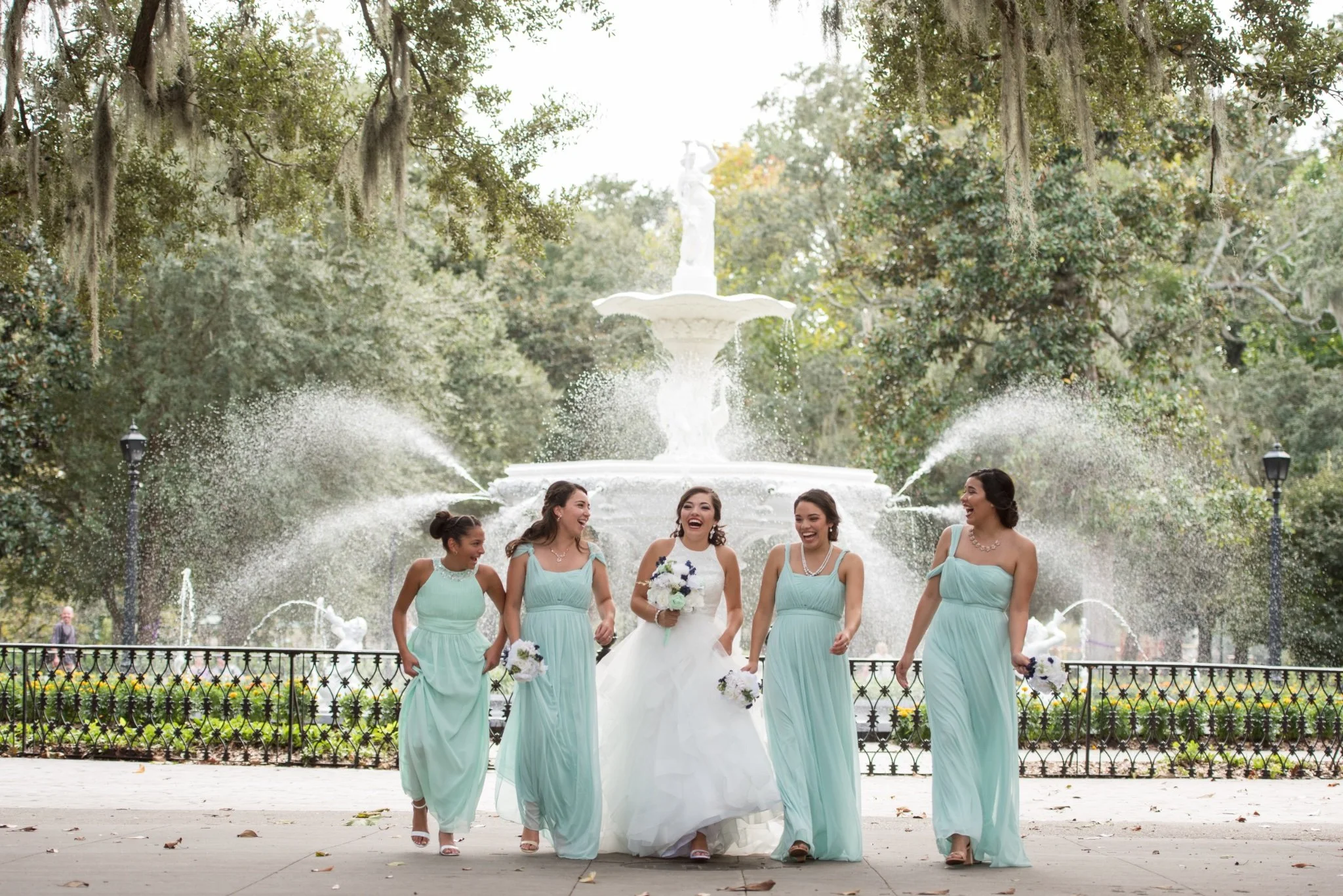 A bride and four bridesmaids walking together and laughing in front of a fountain at a park.