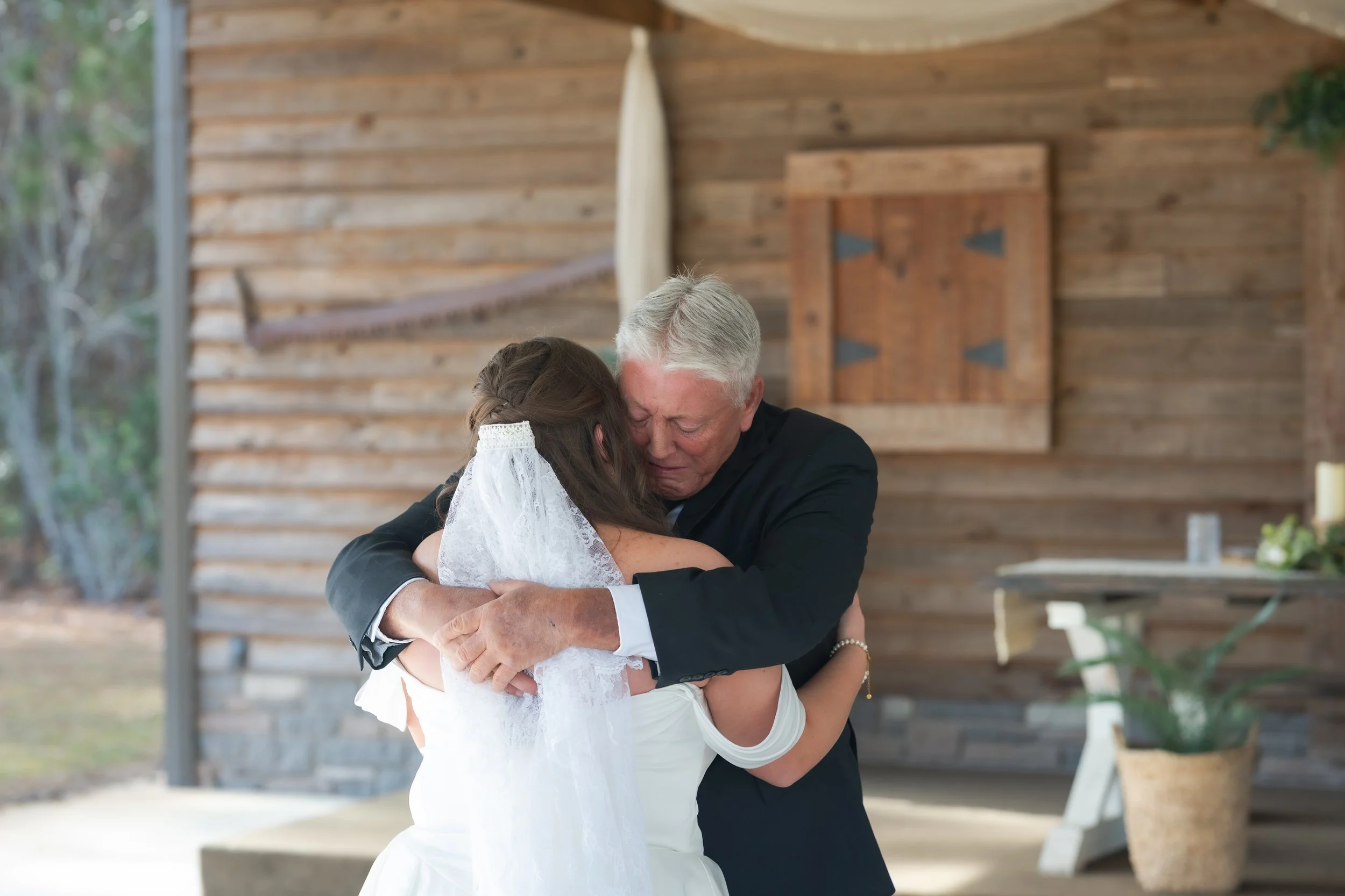 An elderly man in a black suit hugs a woman in a white wedding dress and veil, emotional moment during a wedding ceremony in a rustic setting with wooden walls and greenery outside.