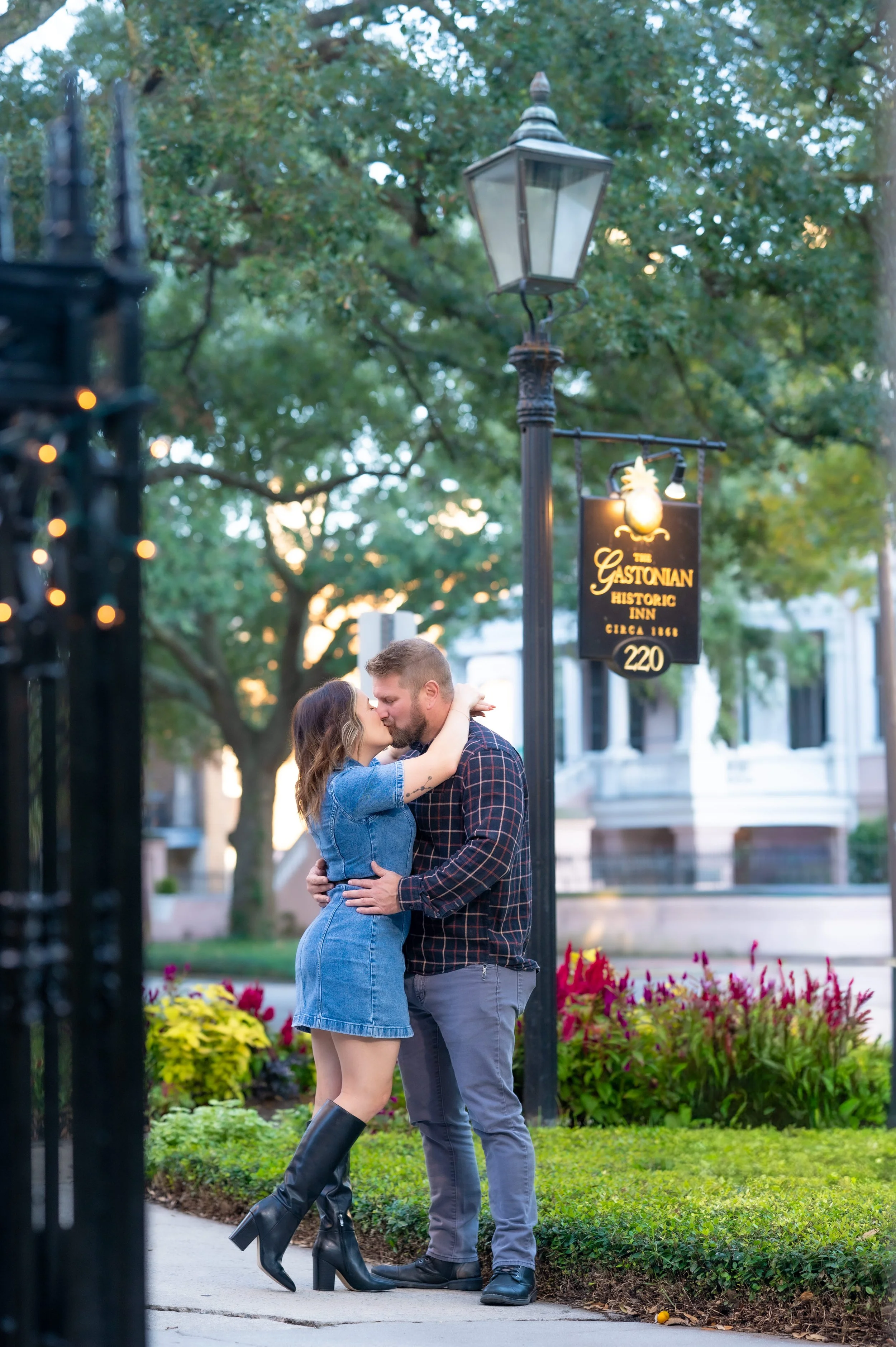 A couple kissing outdoors near a vintage gas lamp and a sign for The Gastonian Historic Inn, with trees, flowers, and a historic building in the background.