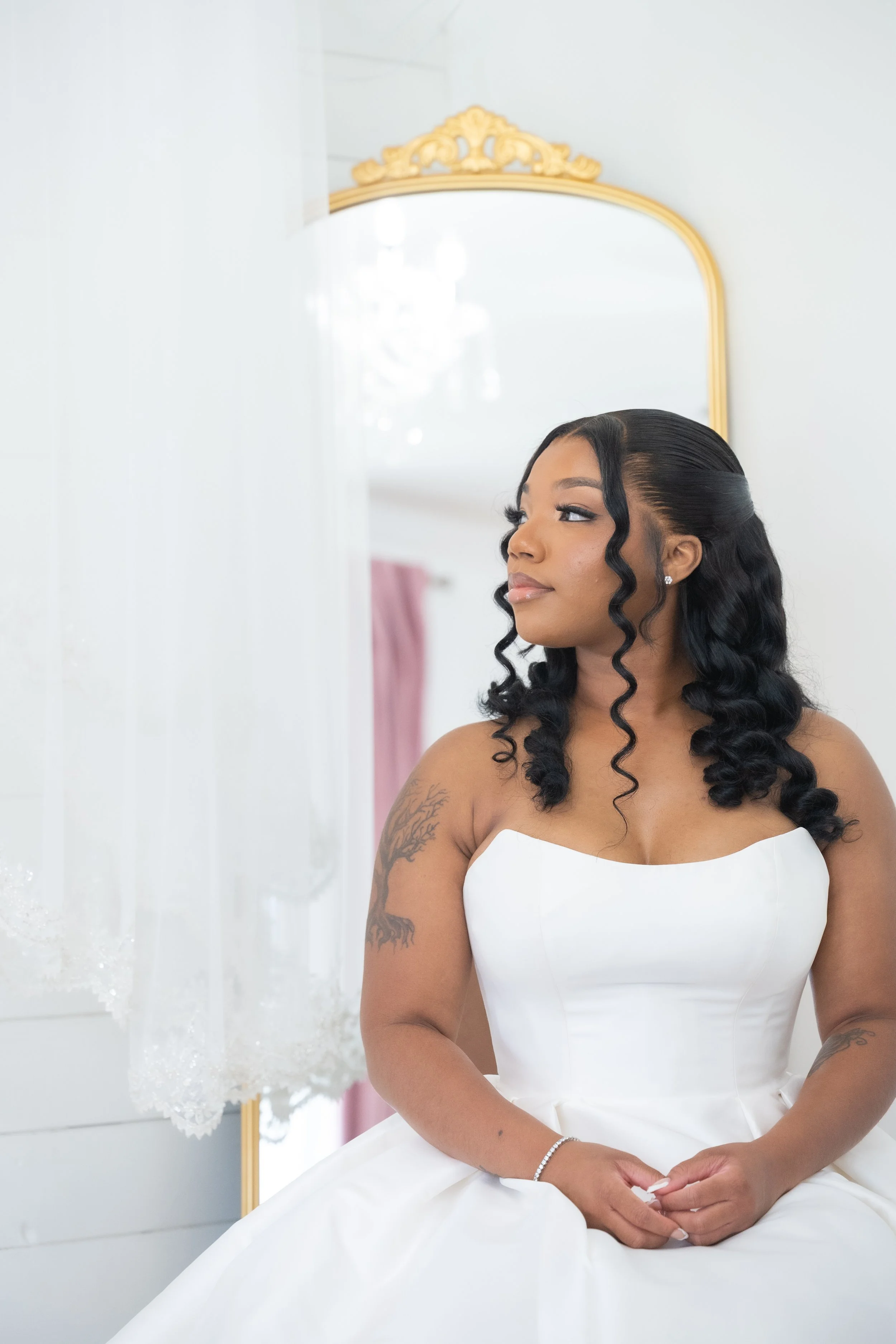 A woman with black wavy hair styled in ringlets, wearing a white strapless dress, sitting in front of a mirror with a gold frame, in a bright room with white walls and pink curtains.