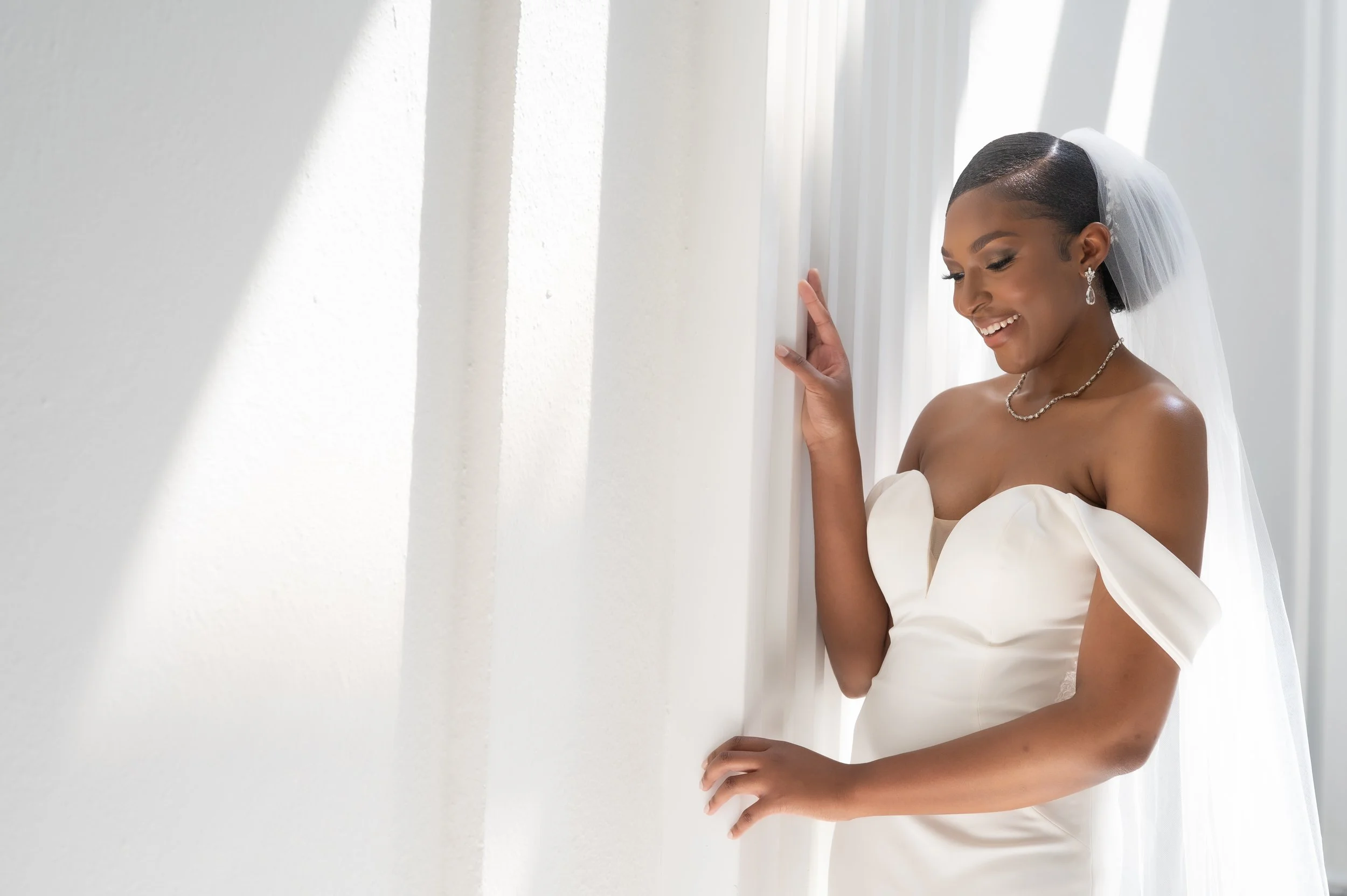 A bride in a white wedding dress with off-shoulder sleeves, smiling and looking down, standing next to a white wall with curtains behind her.