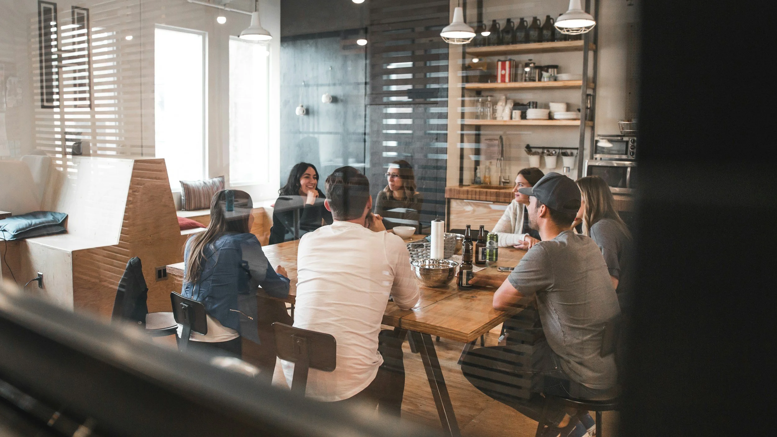 Group of friends sitting around a wooden table in a modern cafe, engaging in conversation and smiling, with beverages and snacks on the table.