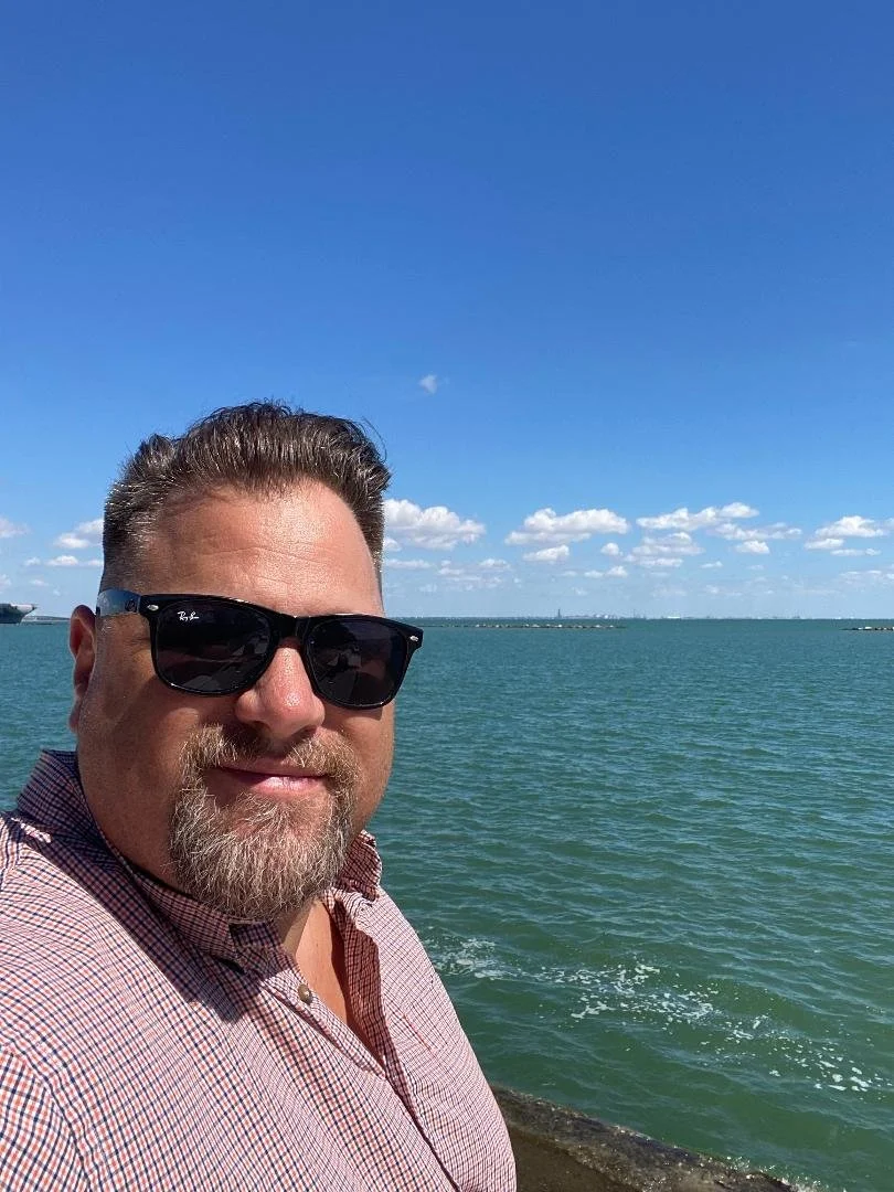 A man with sunglasses and a beard taking a selfie by a body of water on a sunny day with a blue sky and scattered clouds.