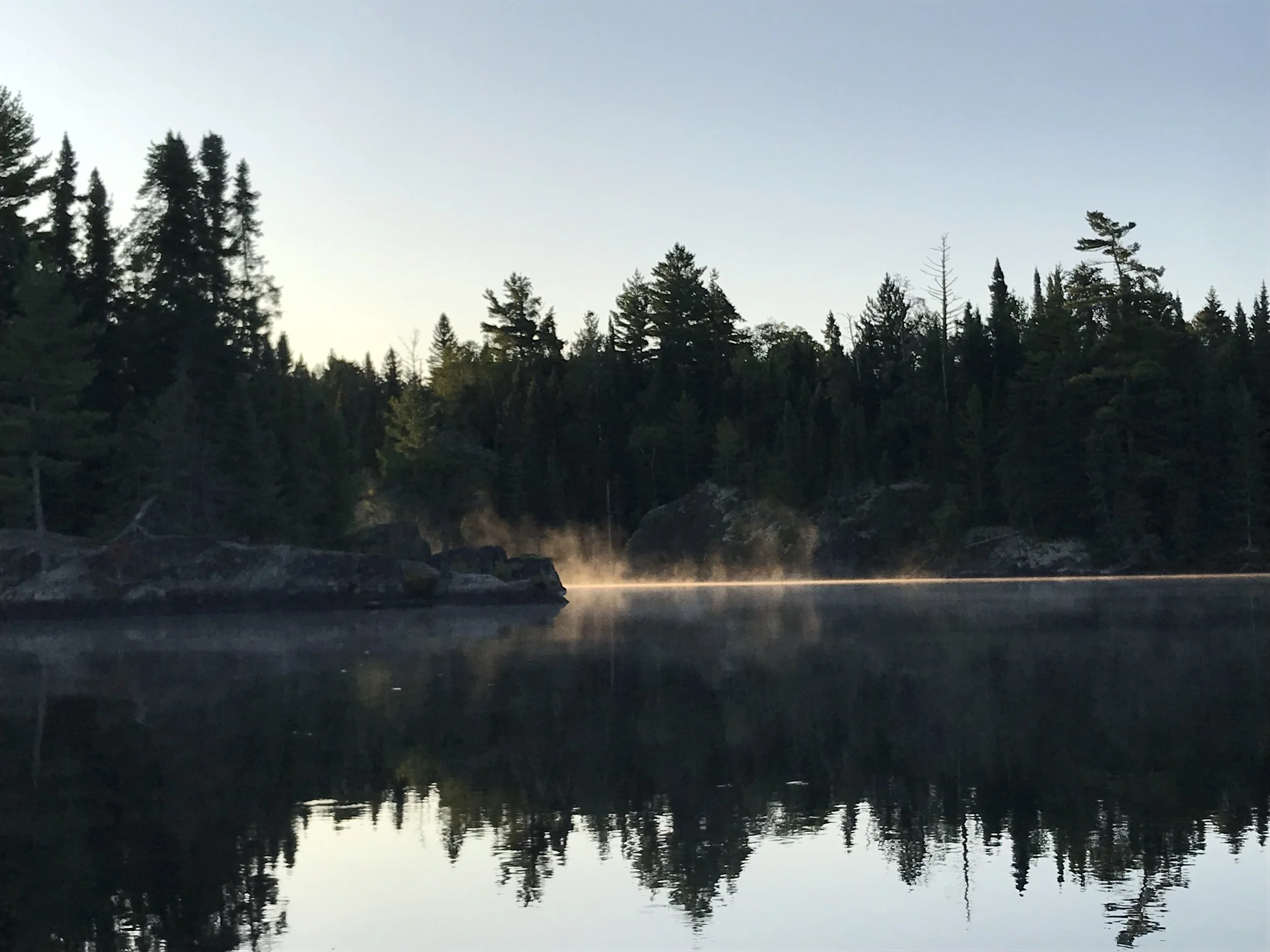 A serene lake surrounded by dense evergreen trees with steam rising from the water's surface at dawn or dusk.