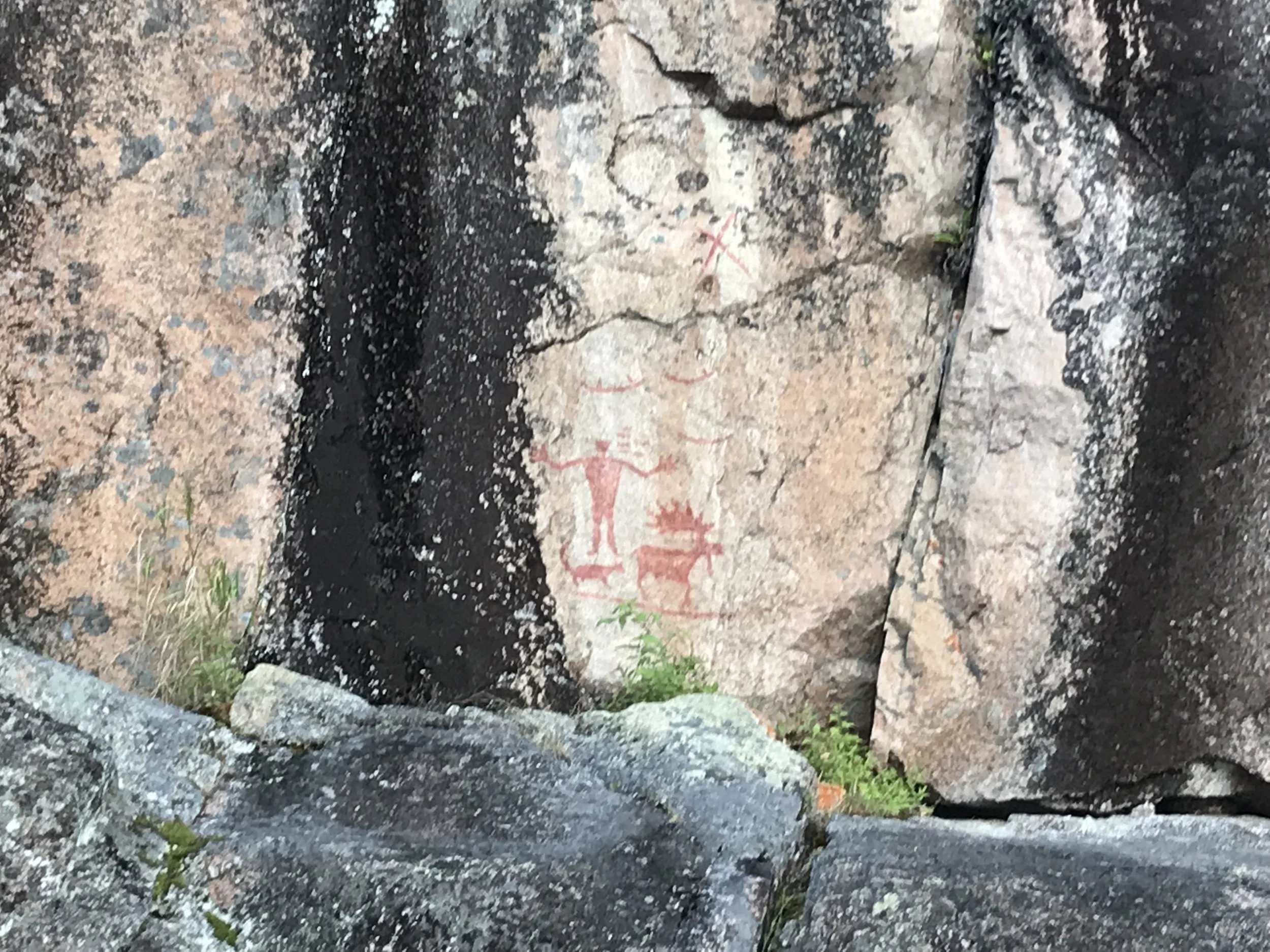 Ancient petroglyphs carved into a rock surface, depicting humanoid figures and a structure, with some red paint markings.