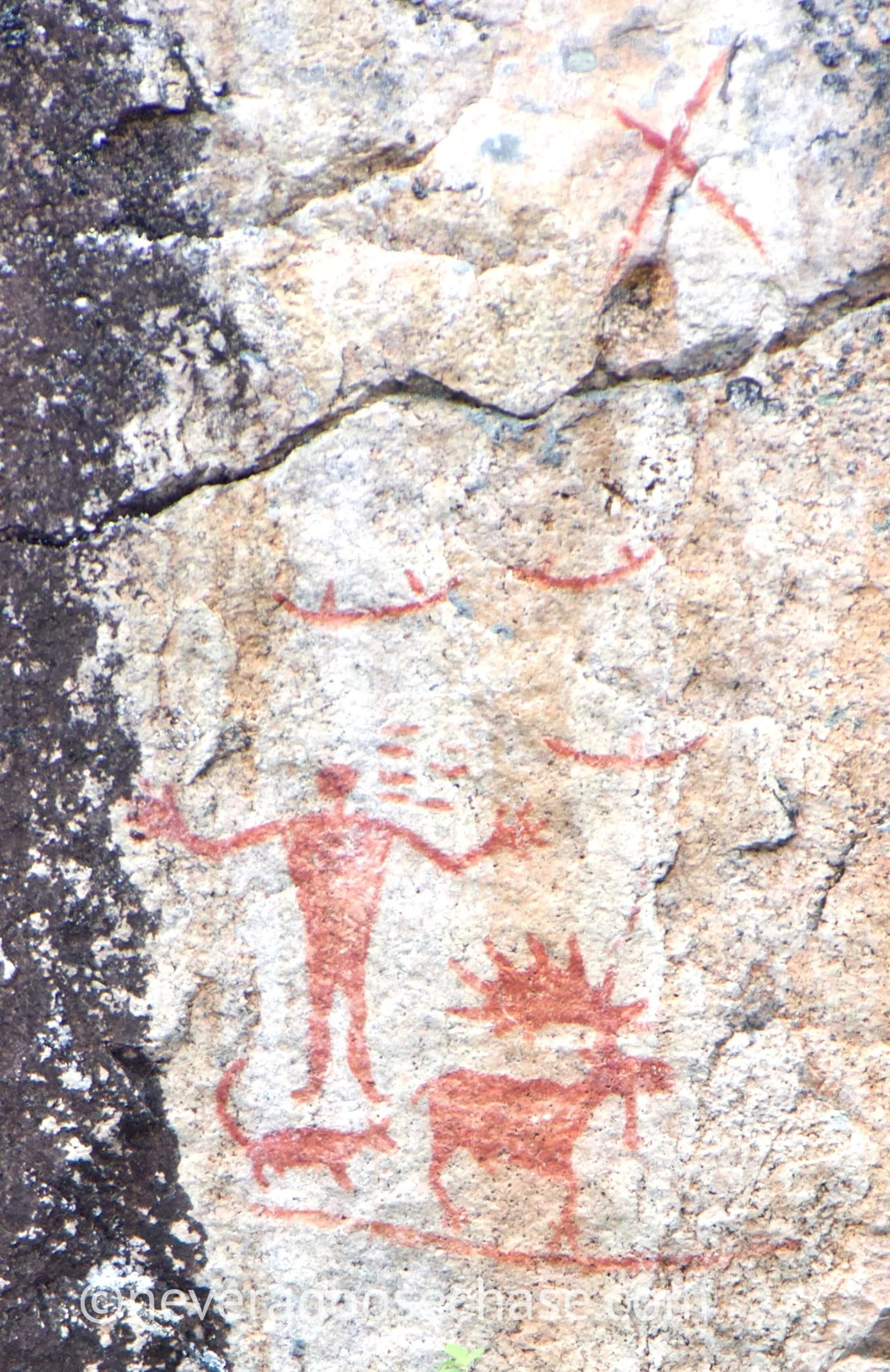 Ancient cave paintings of a human figure with a deer, a bird, and a smiling face, created with reddish pigment on a rocky wall.