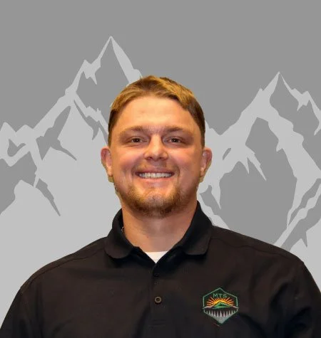 A young man with short, light brown hair and a beard, smiling, wearing a black polo shirt with a mountain logo on the chest, standing in front of a gray background with white mountain outlines.