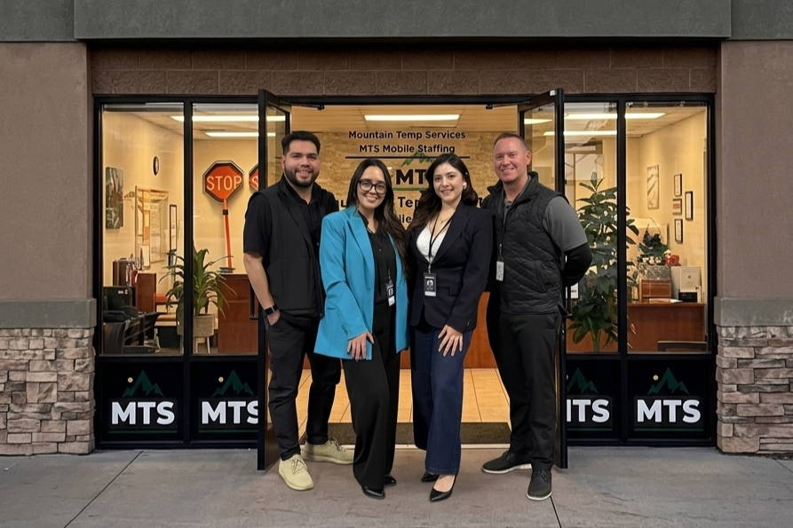 Four people standing outside the entrance of an office building with glass doors, visible indoor plants, and the sign 'Mountain Temp Services MTS Mobile Staffing' in the background.