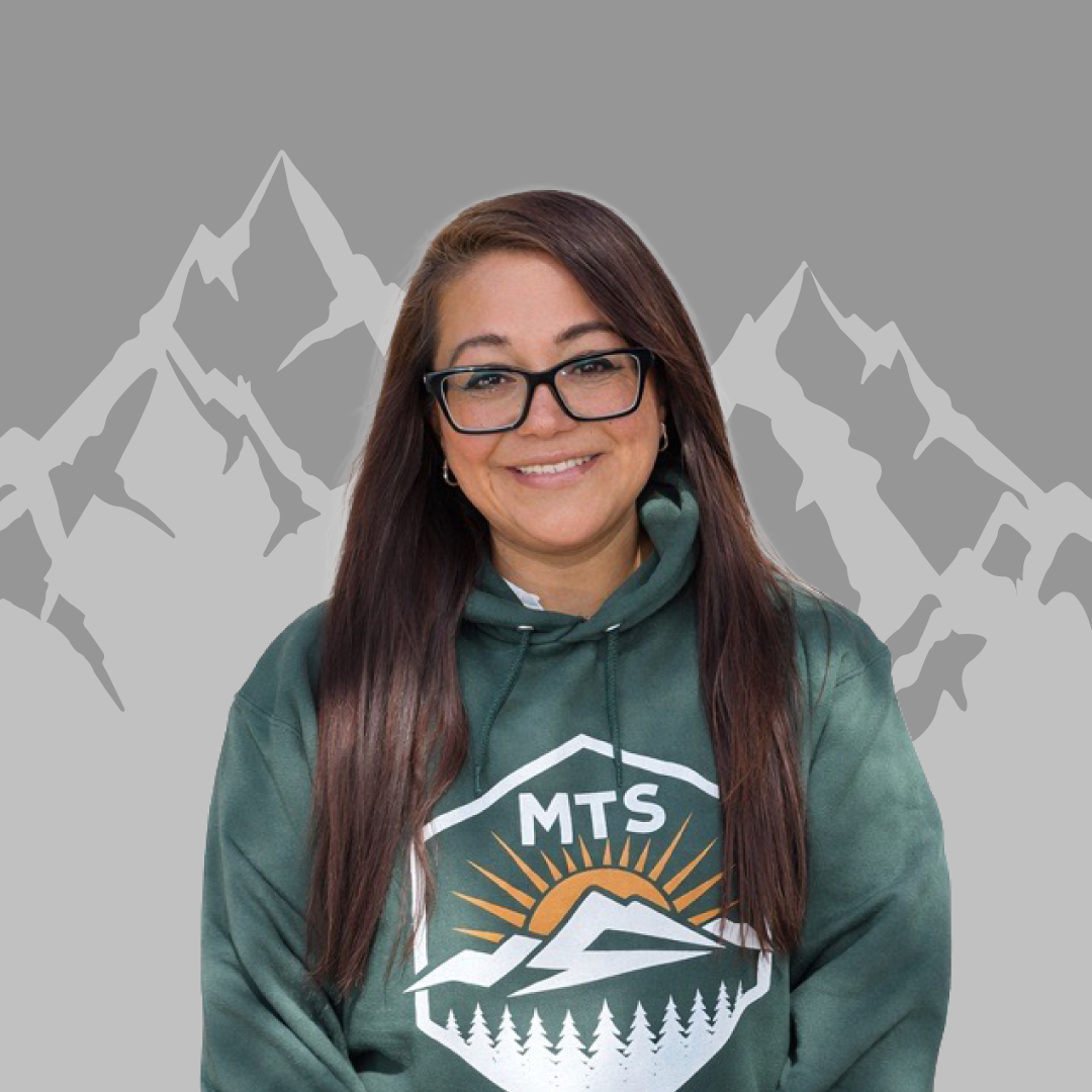 A woman with long brown hair, glasses, and earrings smiling, wearing a green hoodie with a mountain logo and the letters 'MTS' in front of a mountain landscape background.