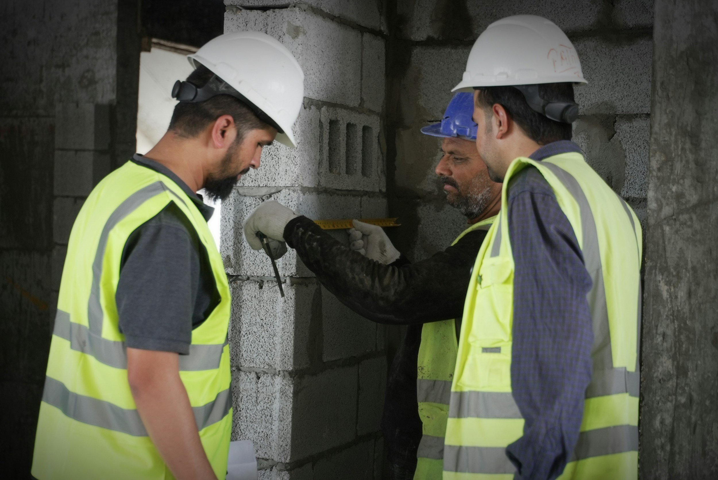 Three construction workers wearing hard hats and safety vests inspecting a wall at a construction site. One worker is measuring the wall with a tape measure.