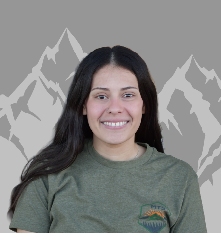 Smiling woman with long dark hair wearing a green shirt with a logo, standing against a gray background with mountain illustrations.