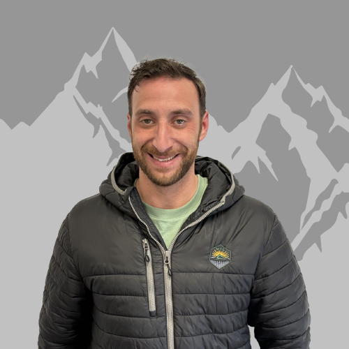 A smiling man with short brown hair and a beard, wearing a black puffer jacket and a green T-shirt, standing in front of a gray background with mountain graphics.