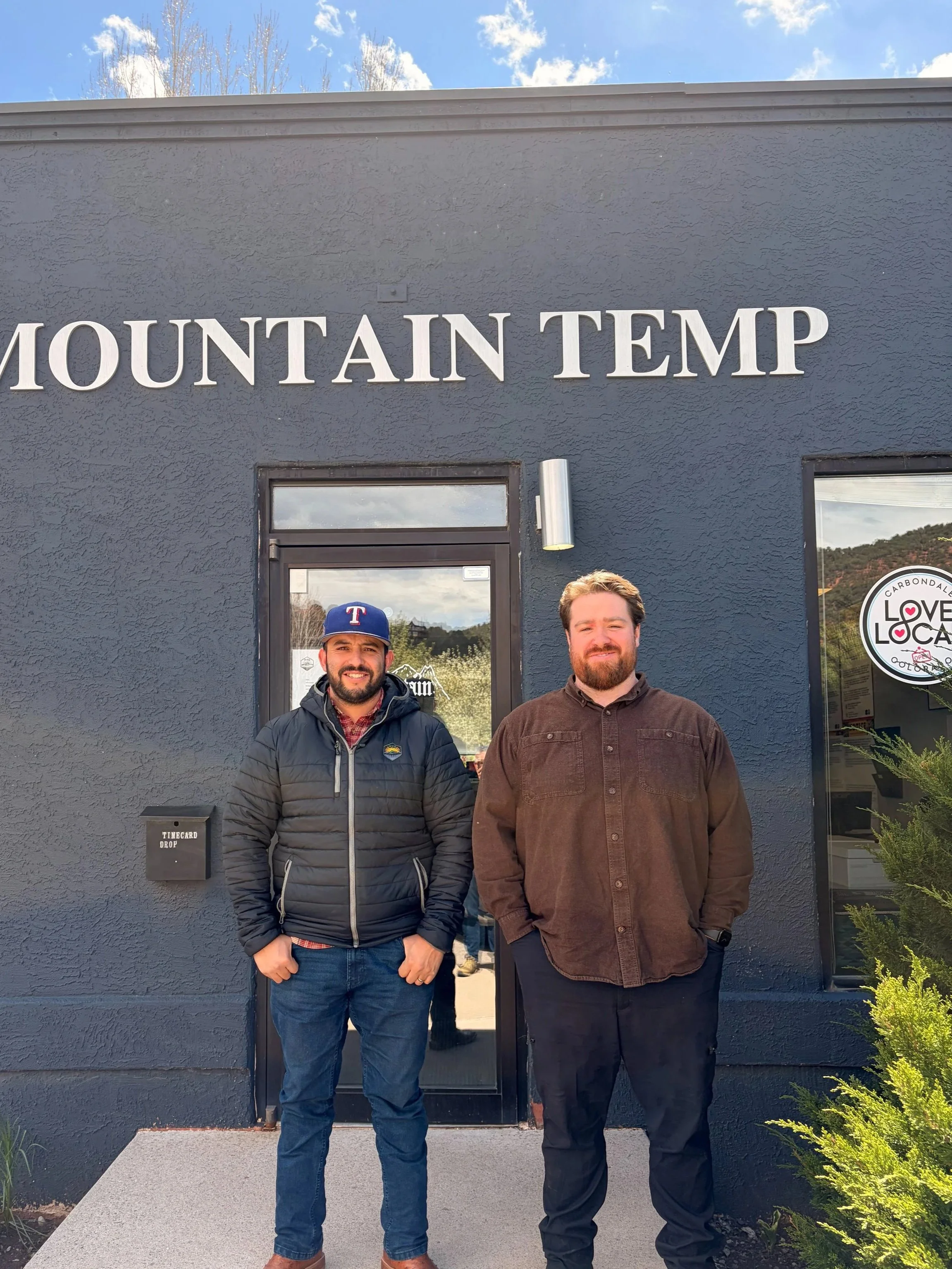 Two men standing in front of a building with the sign 'Mountain Temp'; one is wearing a Texas Rangers cap and a black jacket, the other is in a brown jacket; trees and a blue sky with clouds are visible.