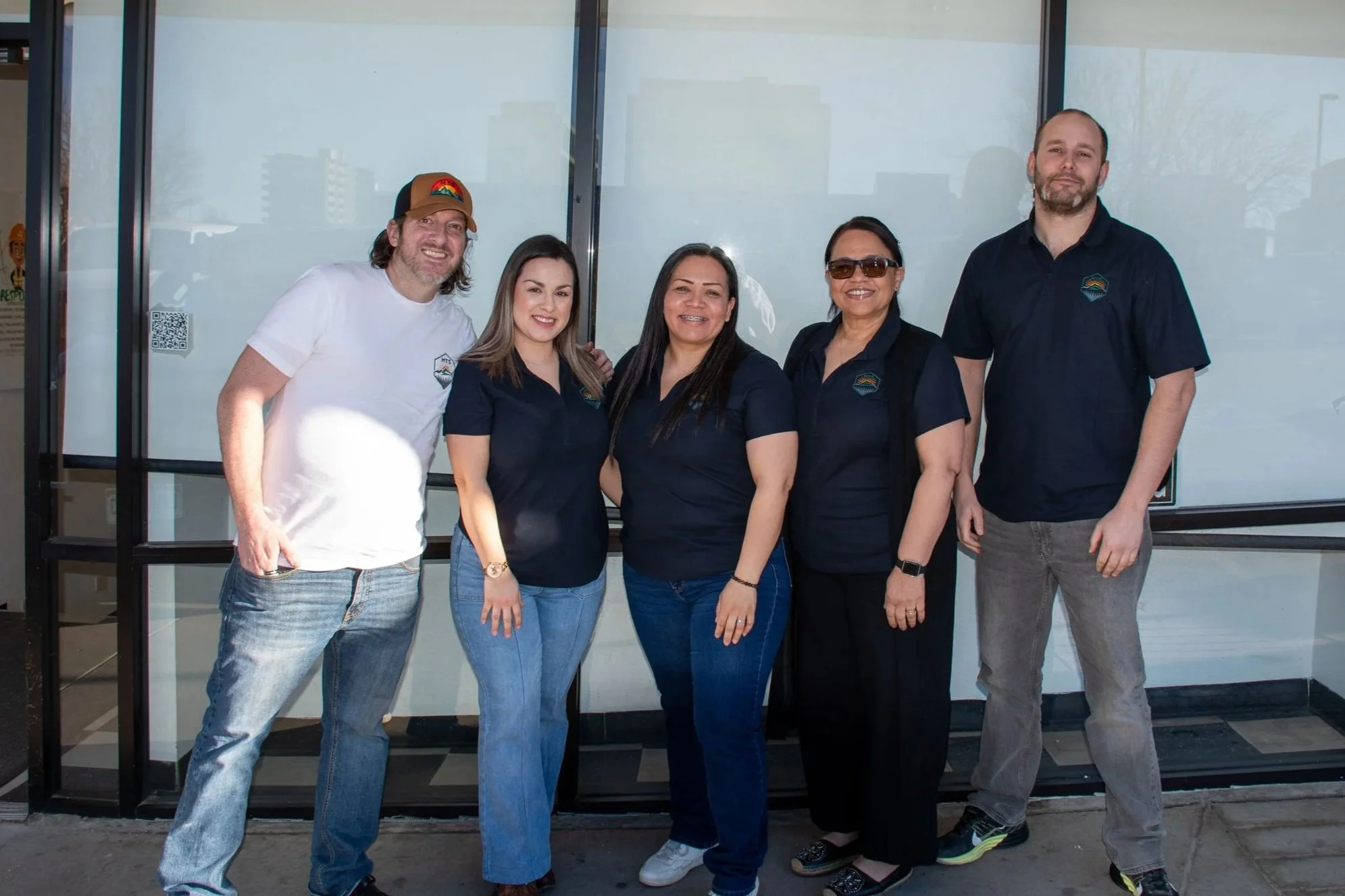 Group of five people standing in front of a glass storefront, smiling at the camera.