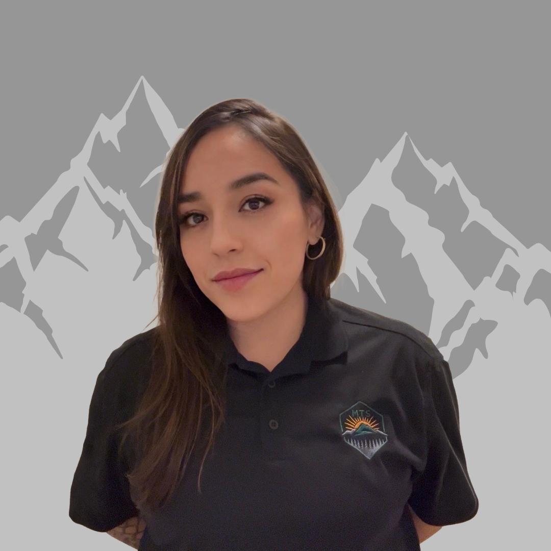 A woman with long brown hair and hoop earrings standing in front of a stylized mountain graphic background, wearing a black polo shirt with a logo that says "MTS" and features a mountain, sun, and water elements.
