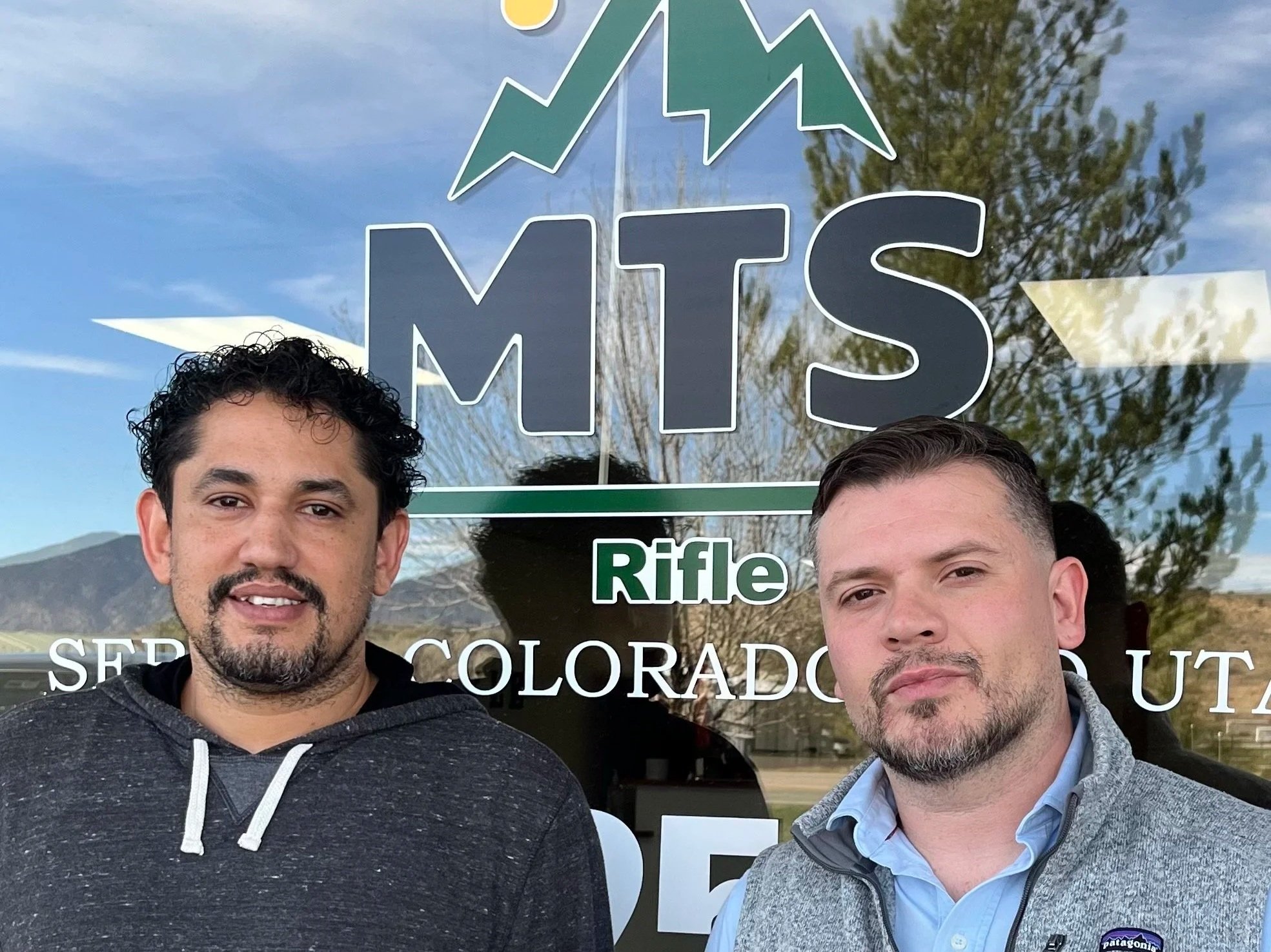 Two men standing in front of a glass window with the MTS logo, mountains, trees, and a partly cloudy sky reflected in the glass.