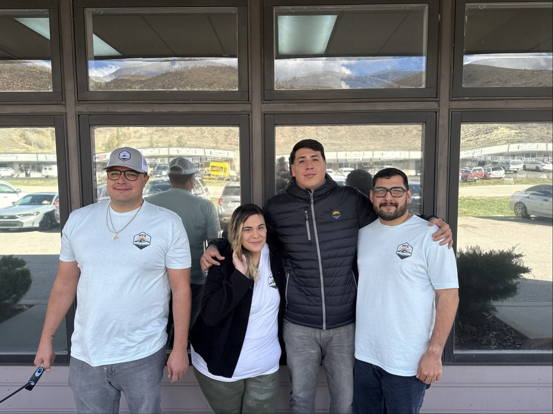 Four people standing together outside a building with large windows and scenic mountain views in the background.
